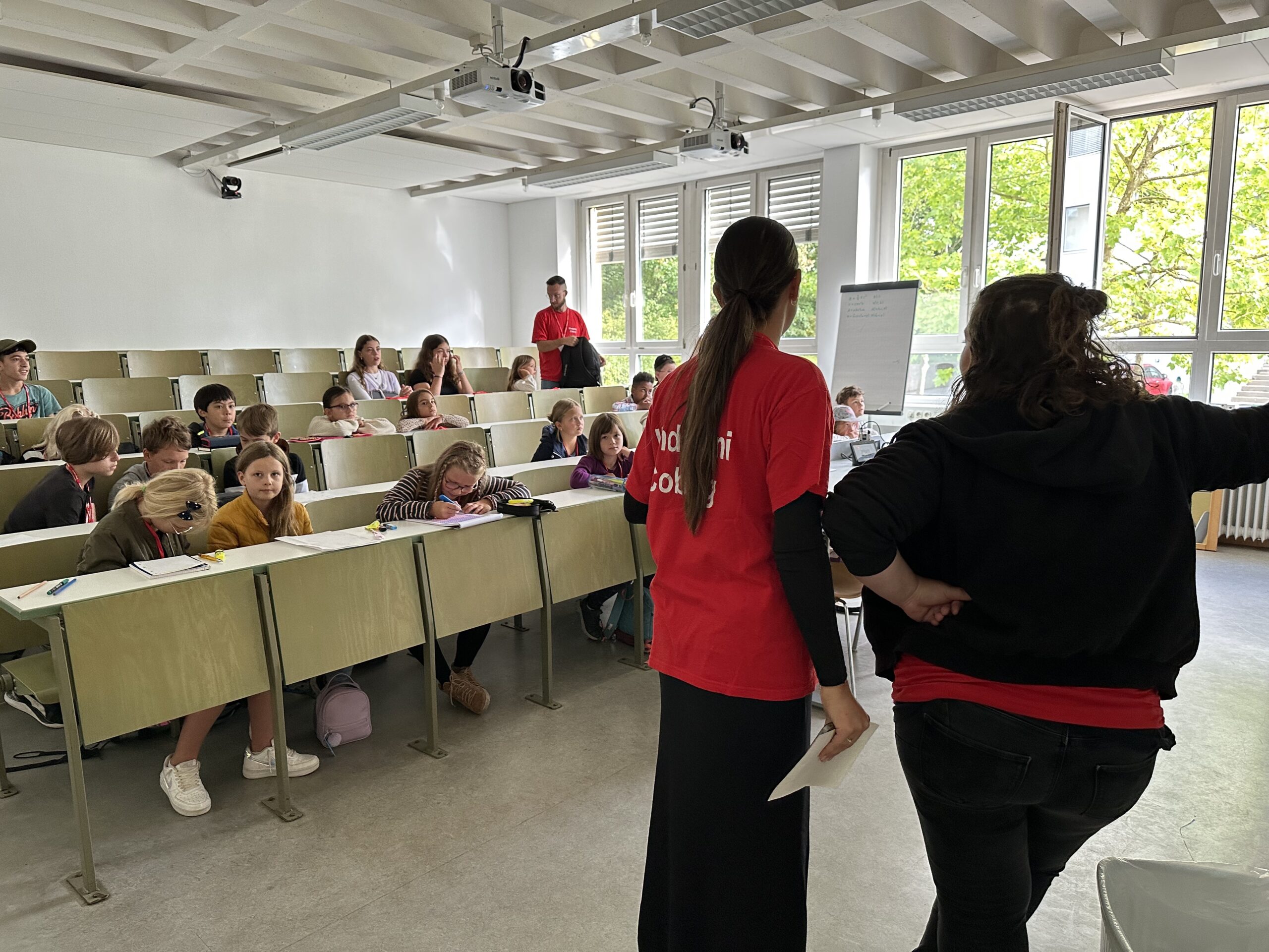 Ein Unterrichtsraum an der Hochschule Coburg ist voll mit Studenten, die in abgestuften Reihen sitzen und sich mit zwei Dozenten in der ersten Reihe unterhalten. Einer deutet auf ein Flipchart, der andere hält ein Blatt Papier, während natürliches Licht durch die großen Fenster fällt.