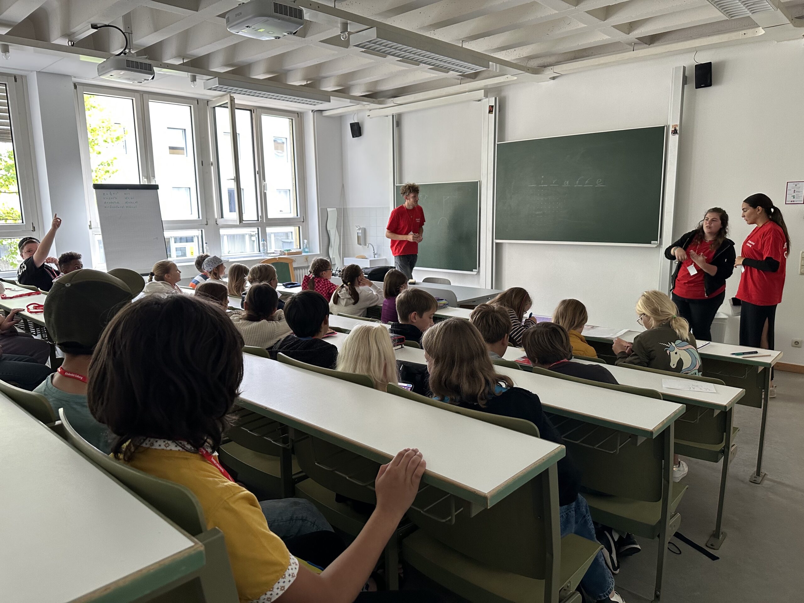 In einem Hörsaal der Hochschule Coburg sitzen die Studenten an mehreren Tischen und unterhalten sich mit einem Lehrer und zwei Assistenten in roten Hemden, die an der Tafel stehen. Die großen Fenster des Raums und das Neonlicht erhellen die eifrigen Gesichter der Studenten mit erhobenen Händen.