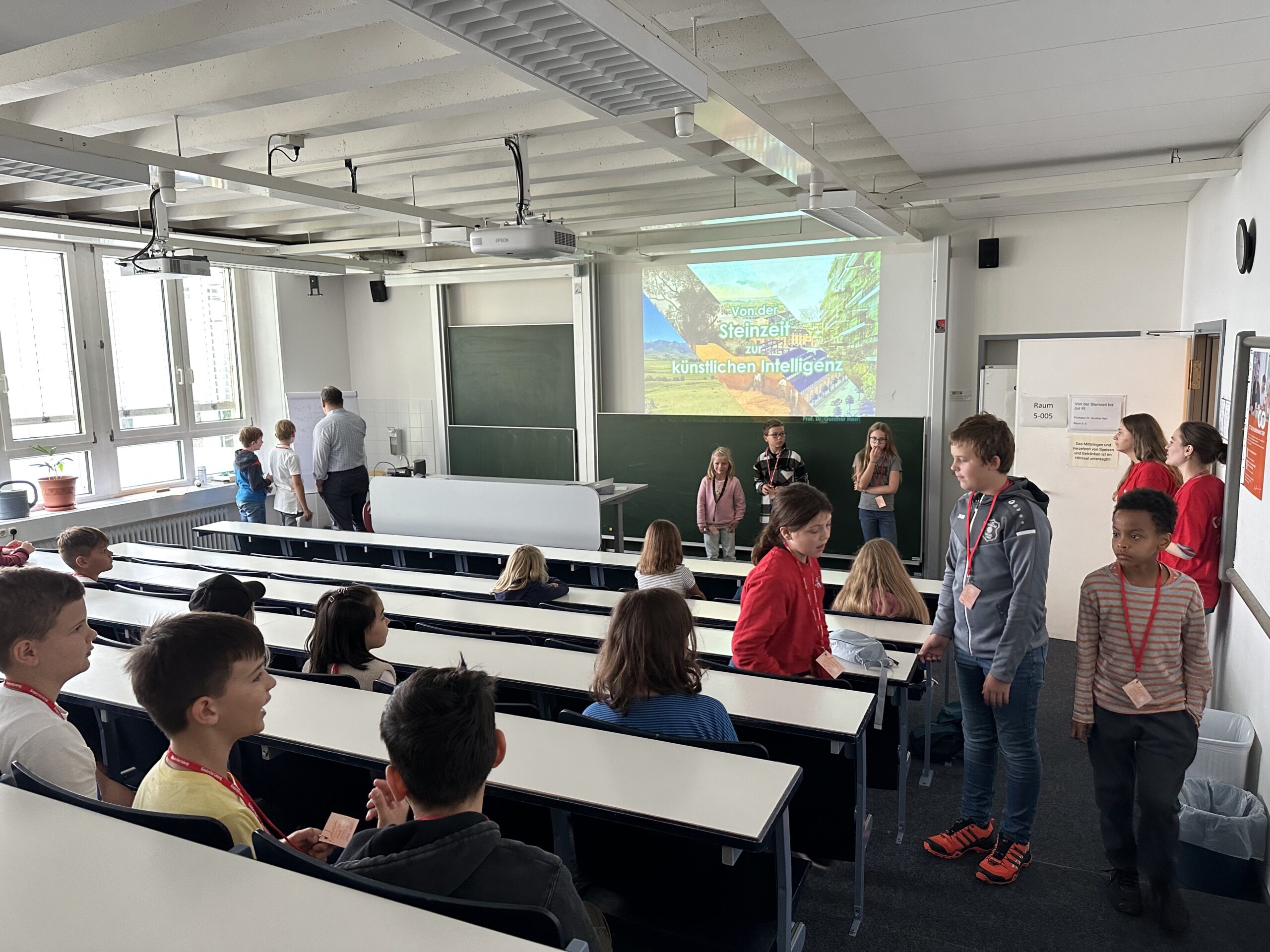 Eine Gruppe von Kindern und ein paar Erwachsenen versammelt sich in einem Klassenzimmer mit Sitzreihen. Einige Schüler stehen vorne, wo „Steinerlein“ auf eine Leinwand projiziert wird, was die pädagogische Atmosphäre der Hochschule Coburg widerspiegelt. Andere sitzen, während ein Erwachsener an der Tafel steht.