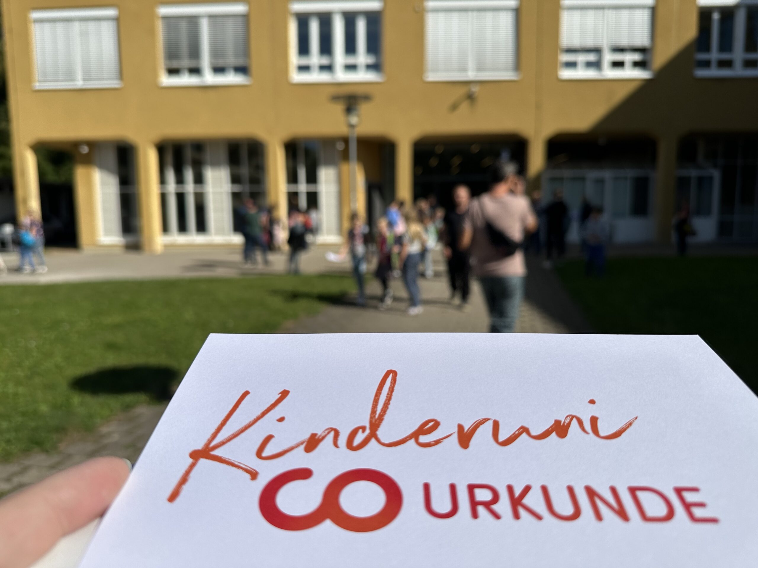 Eine Hand hält ein „Kinderuni-Urkunde“-Zertifikat. Im Hintergrund wimmelt es von Kindern und Erwachsenen der Hochschule Coburg.