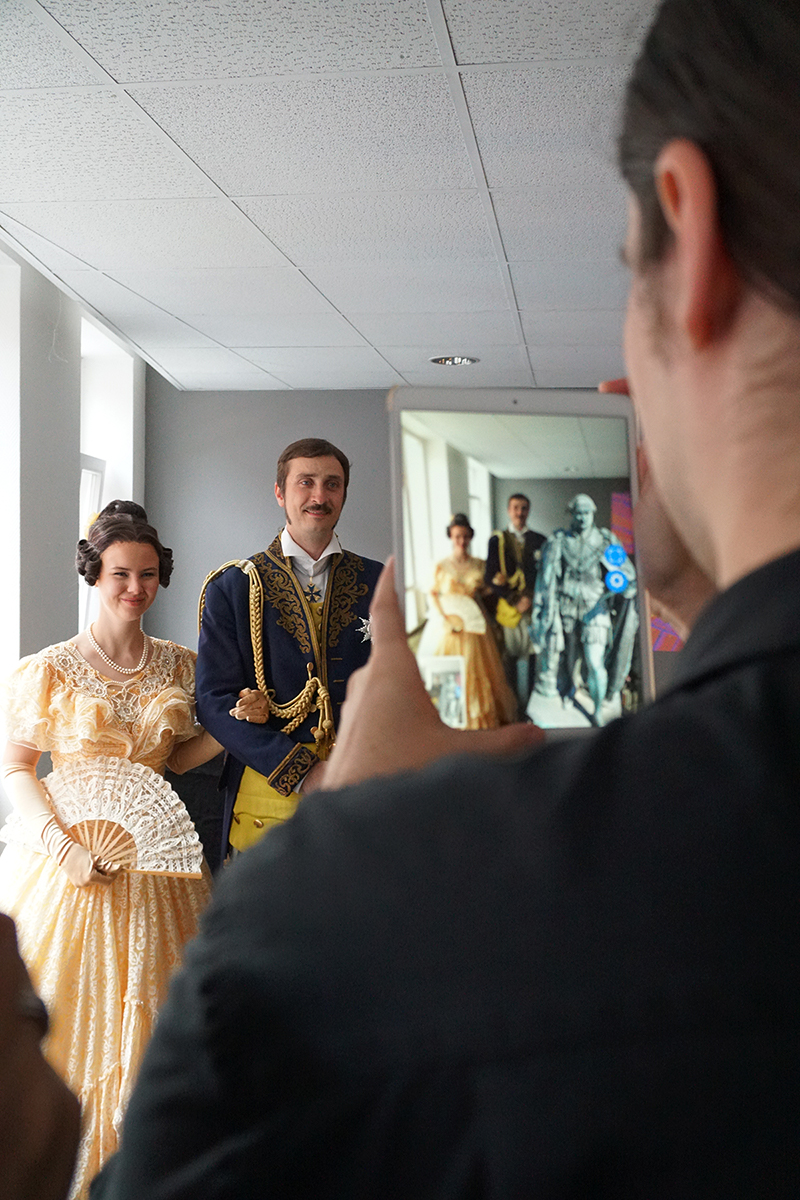 Eine Person macht ein Foto von einem lächelnden Paar, das elegant in historischer Kleidung gekleidet ist, an der Hochschule Coburg. Die Frau hält einen Spitzenfächer und trägt ein gelbes Kleid, während der Mann einen verzierten blauen Mantel trägt. Ein Tablet zeigt das gespiegelte Bild dieser bezaubernden Szene.