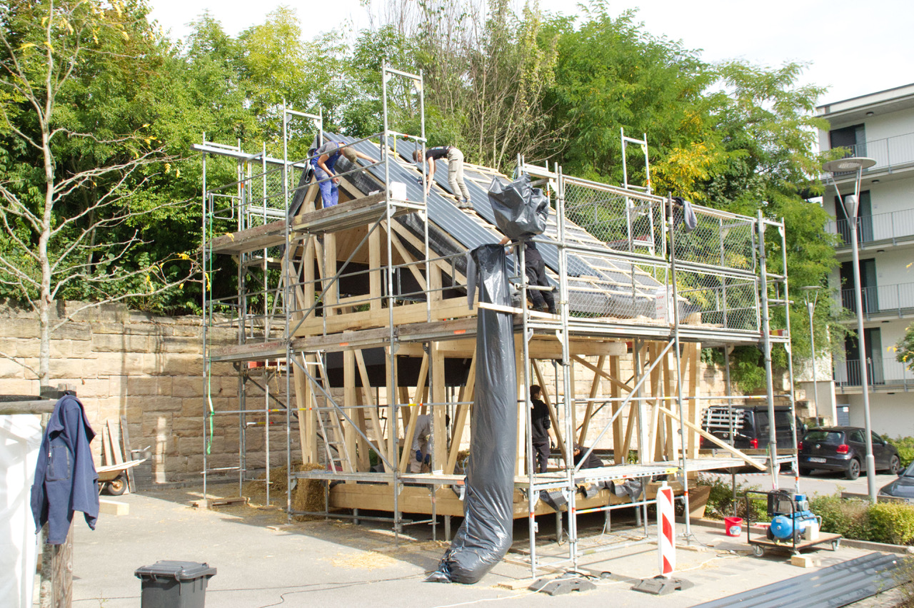 Ein im Bau befindliches Fachwerkhaus, umgeben von einem Gerüst, spiegelt die Architekturstudien der Hochschule Coburg wider. Arbeiter montieren Dachplatten inmitten von überall verstreuten Materialien und Werkzeugen. Neben einer Steinmauer und Bäumen sind im Hintergrund nahe gelegene Gebäude zu sehen.