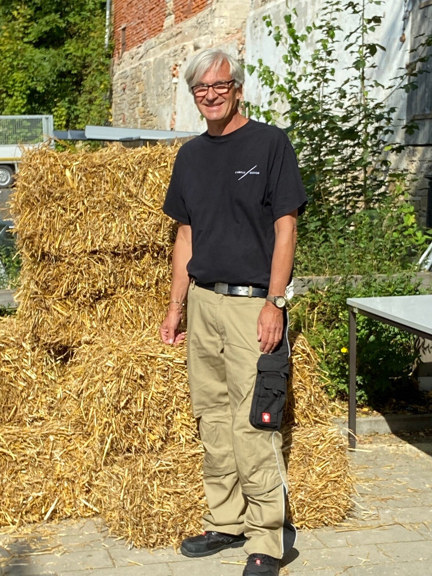 Eine Person mit kurzen grauen Haaren steht im Freien vor gestapelten Heuballen. Sie trägt ein schwarzes T-Shirt, beige Cargohosen und schwarze Schuhe. Dahinter erinnern das Grün und ein Gebäude an die ruhige Umgebung der Hochschule Coburg.