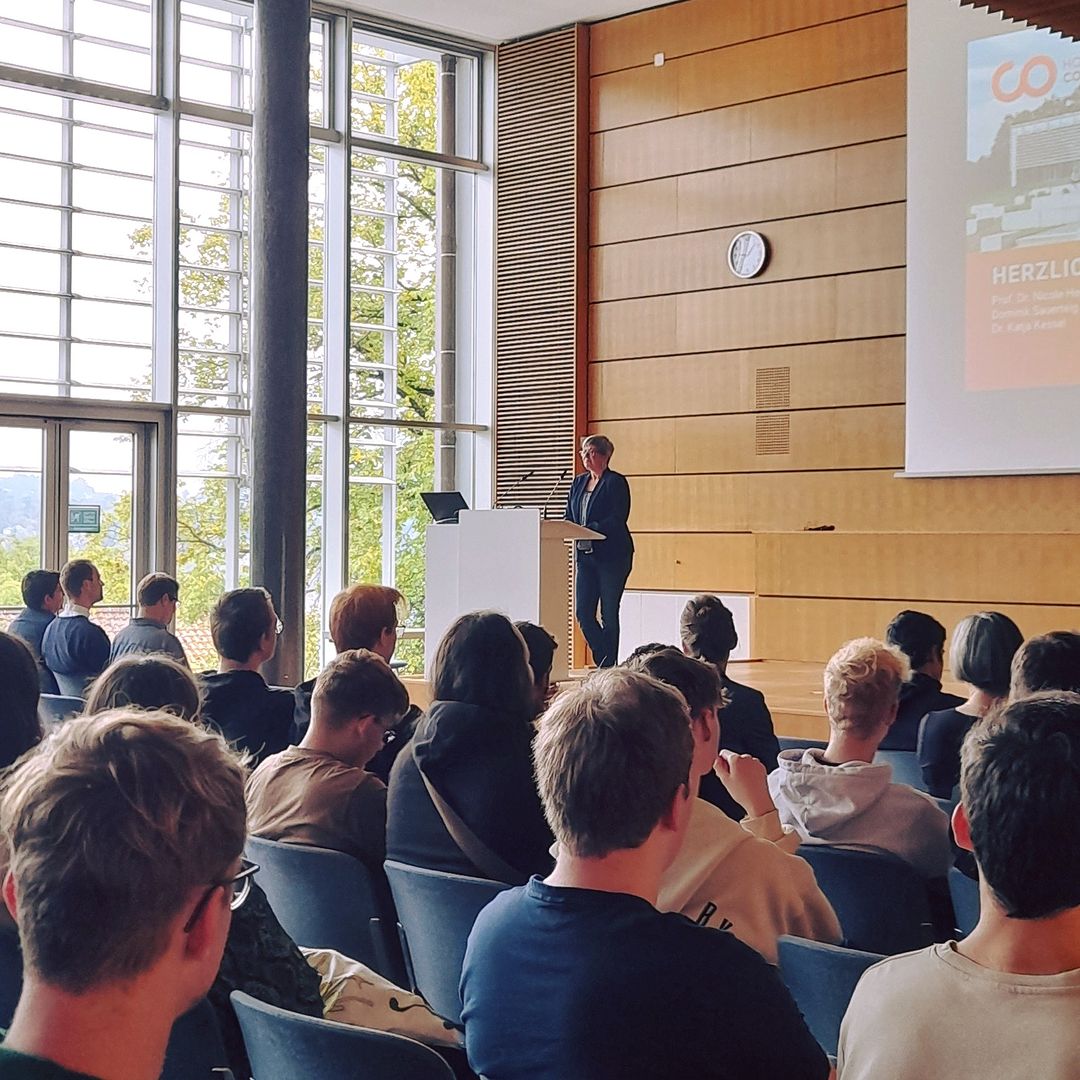 Eine Person steht an einem Rednerpult in einem modernen Hörsaal mit Holzwänden und großem Fenster an der Hochschule Coburg. Sie hält einen Vortrag vor einem sitzenden Publikum, während auf der Leinwand eine Folie angezeigt wird.