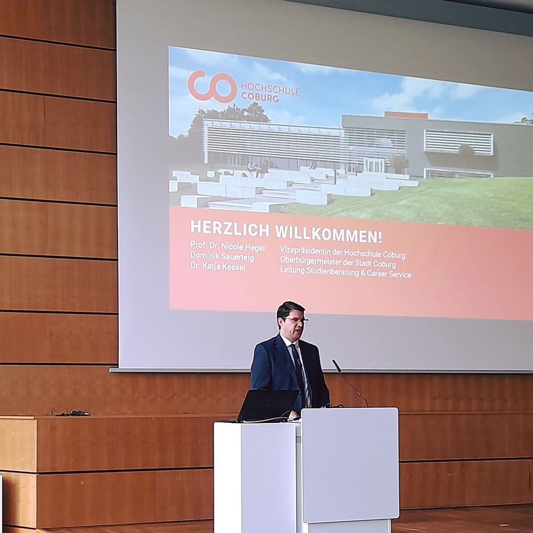 Eine Person steht an einem Podium und hält einen Vortrag in einem Hörsaal der Hochschule Coburg. Dahinter ist auf einem Bildschirm das Logo und „Herzlich Willkommen!“ neben einigen Namen zu sehen. Ergänzt wird die Szene durch ein modernes Gebäude im Hintergrund, das den Innovationsgeist der Hochschule widerspiegelt.