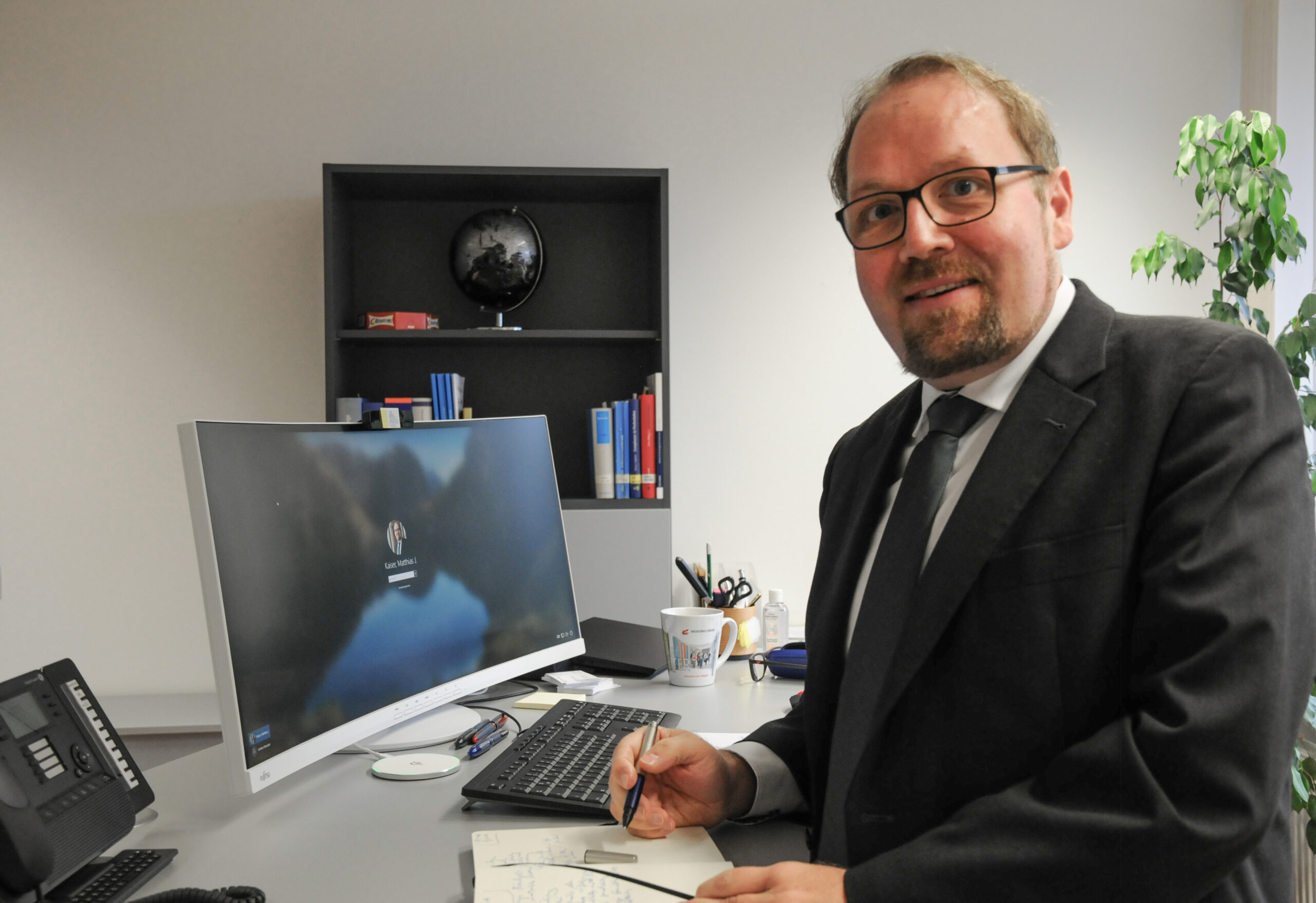 Ein Mann im Anzug steht an einem Schreibtisch mit einem Computermonitor, auf dem der Anmeldebildschirm der Hochschule Coburg angezeigt wird. Er hält einen Stift und ein Notizbuch in der Hand, im Hintergrund stehen Büromaterialien und Bücher auf Regalen. Auf dem Regal steht ein kleiner Globus, der für eine internationale Note sorgt.