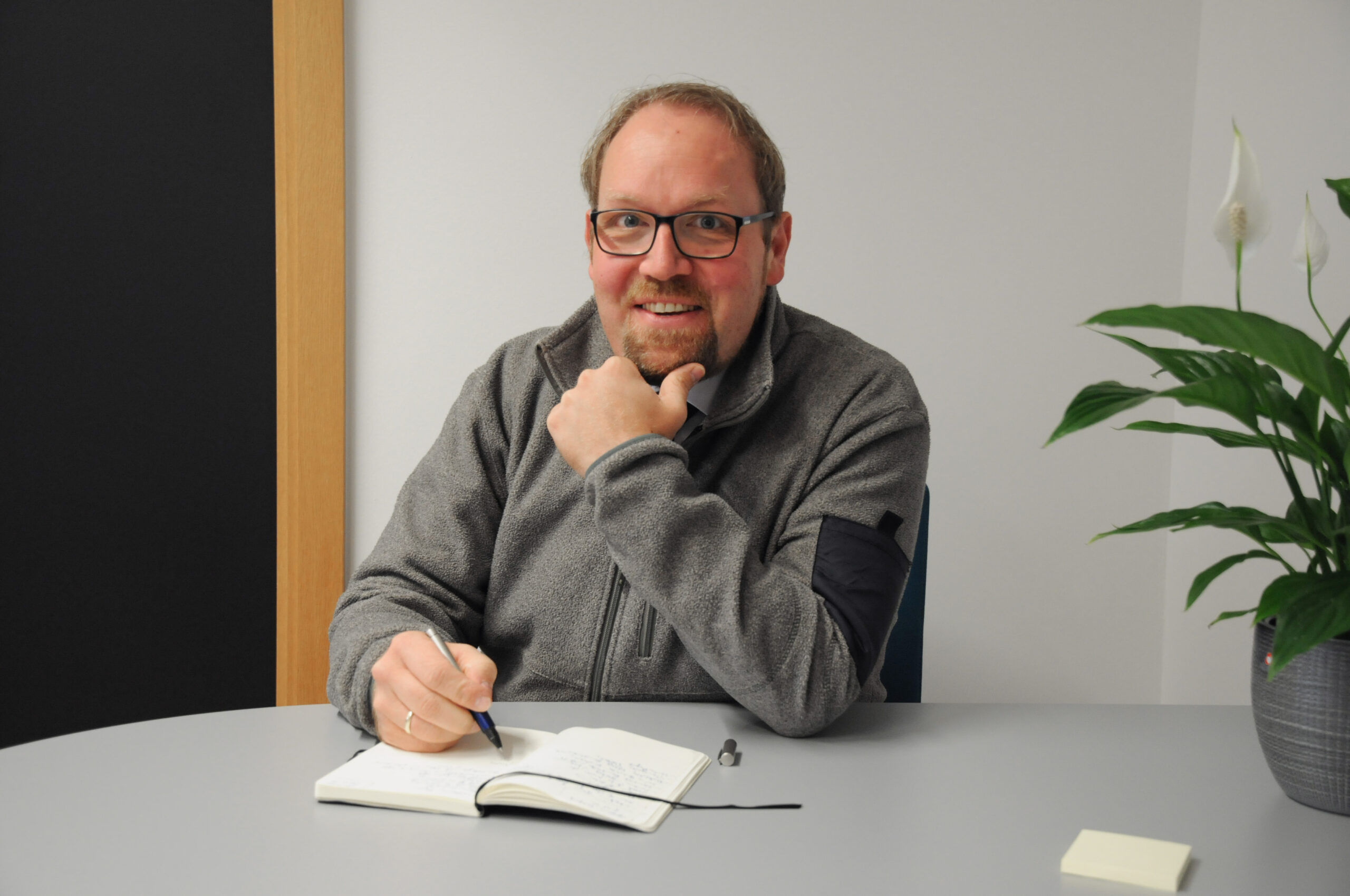 Ein Mann mit Brille und Bart sitzt an einem Tisch und lächelt in die Kamera. Er trägt eine graue Jacke und hält einen Stift über einem offenen Notizbuch mit dem Logo der Hochschule Coburg. Eine Topfpflanze verleiht der Szene einen Hauch von Grün.