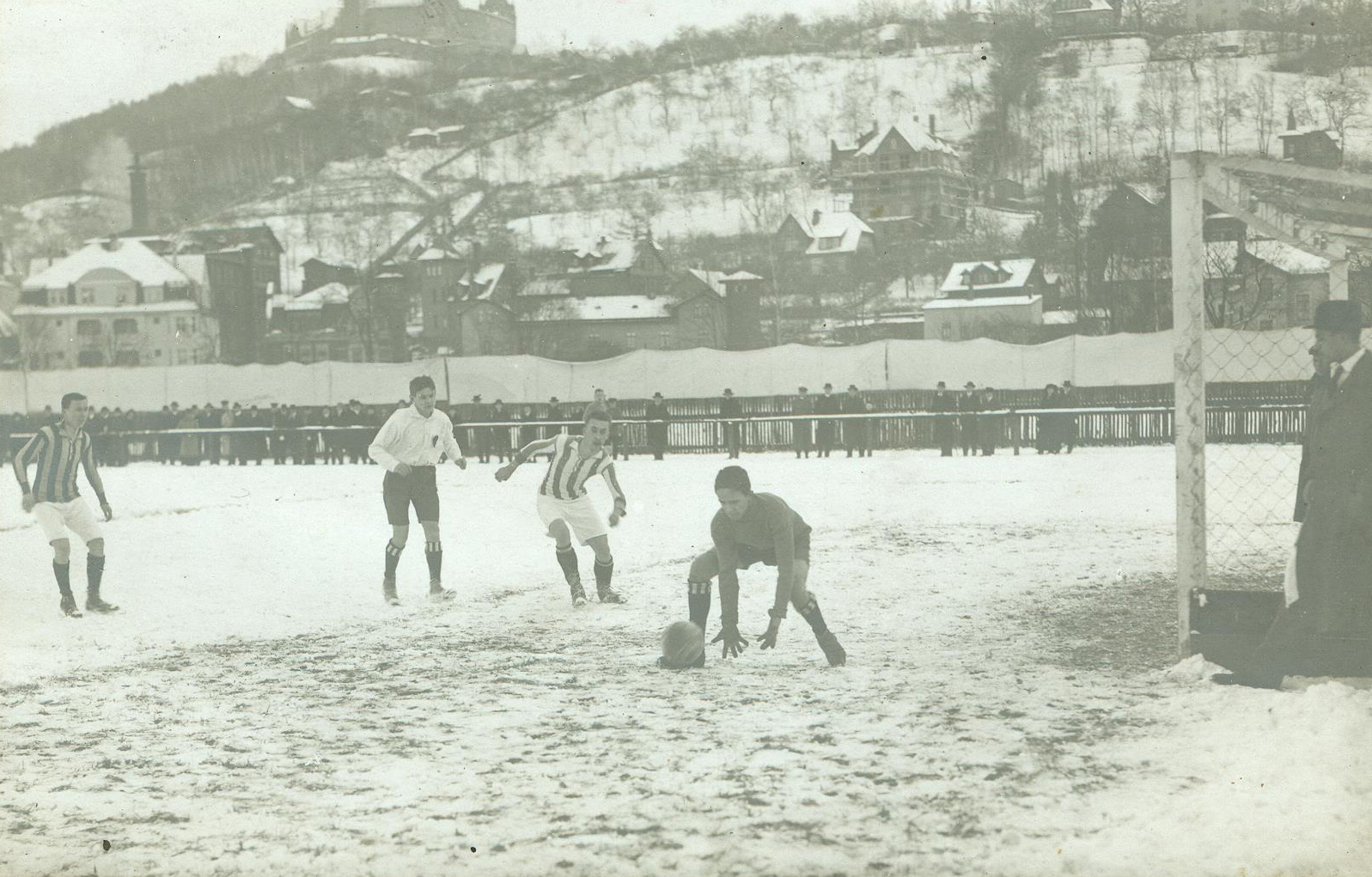 Ein altes Foto zeigt ein Fußballspiel im Schnee in der Nähe der Hochschule Coburg. Der dunkel gekleidete Torwart bückt sich, um den Ball aufzuheben, als Spieler in gestreiften Trikots näher kommen. Hinter ihnen schaut eine Menge gespannt hinter einem Zaun zu, schneebedeckte Hügel rahmen die Szene ein.