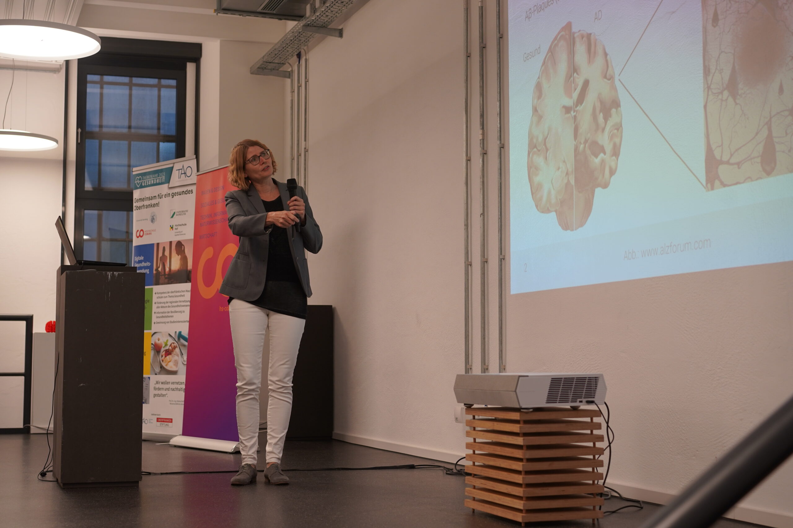 Eine Person steht in einem modernen Konferenzraum der Hochschule Coburg, hält ein Mikrofon in der Hand und präsentiert ein auf eine Leinwand projiziertes Gehirndiagramm. Sie trägt Blazer und Hose, im Hintergrund sind ein Banner und ein Tisch zu sehen.