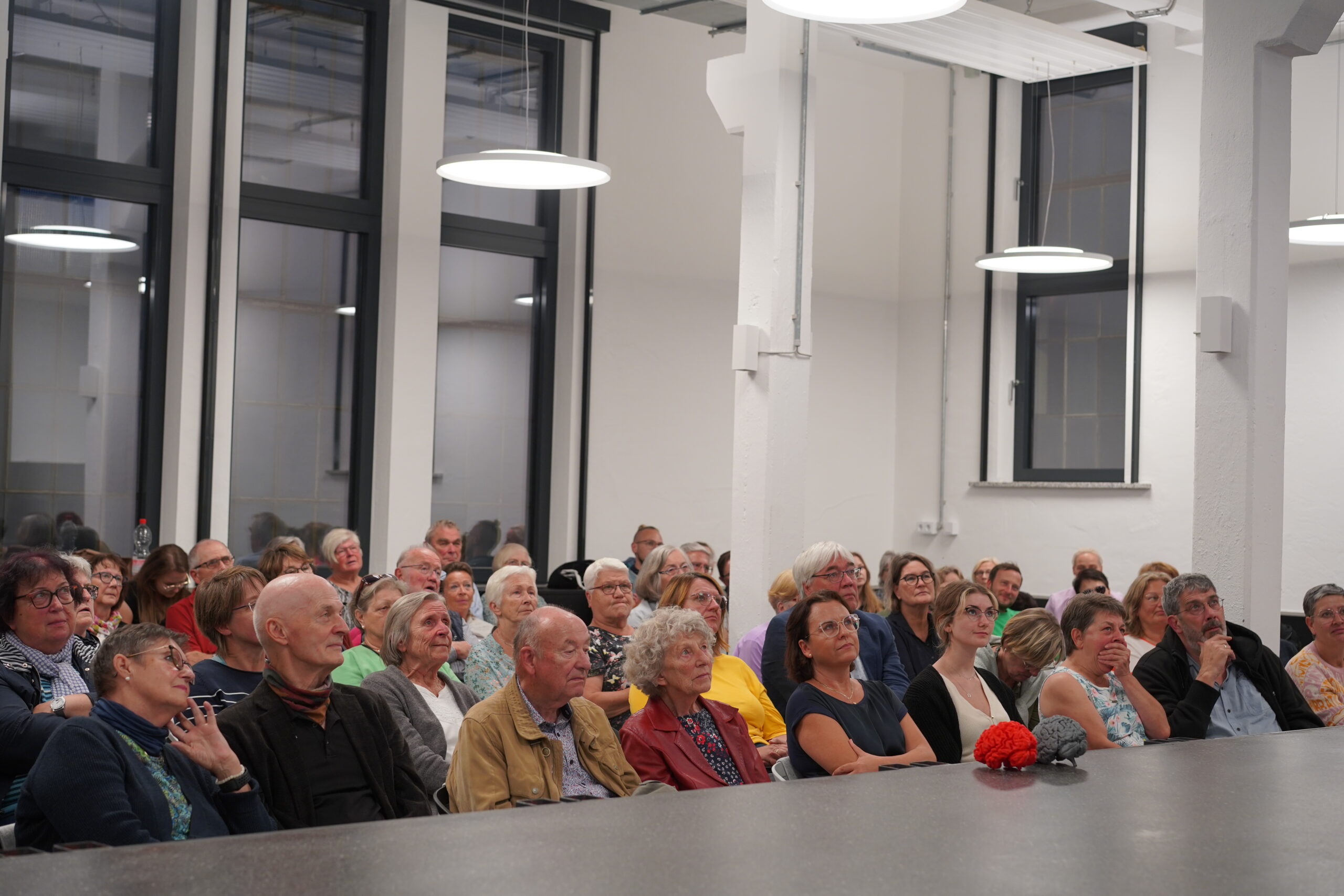 Eine bunt gemischte Gruppe von Menschen sitzt aufmerksam in einem gut beleuchteten Raum mit großen Fenstern und hohen Decken und nimmt an einer Indoor-Veranstaltung der Hochschule Coburg teil.