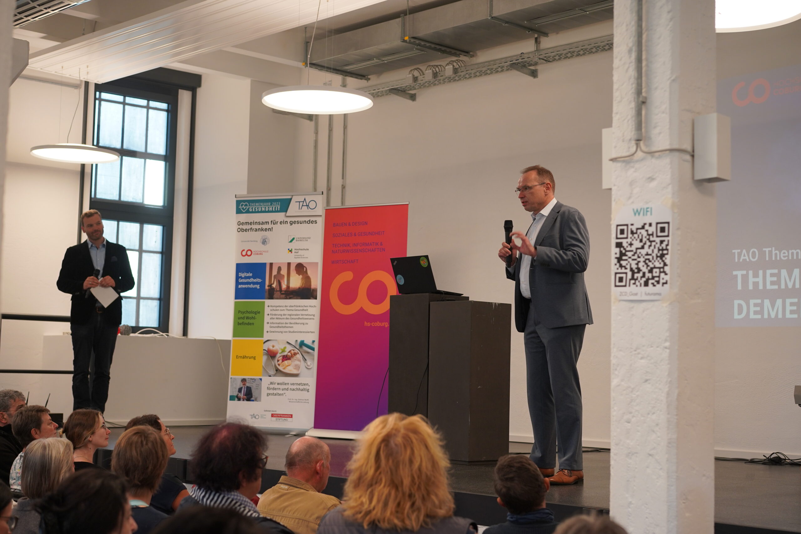 Ein Mann im Anzug spricht auf einem Podium in einem Konferenzraum zu einem Publikum. Hinter ihm repräsentieren ein großer Bildschirm und bunte Banner das Branding der Hochschule Coburg. Daneben hält ein weiterer Mann ein Mikrofon. Auf einer Säule in der Nähe ist gut sichtbar ein QR-Code-Zeichen angebracht.