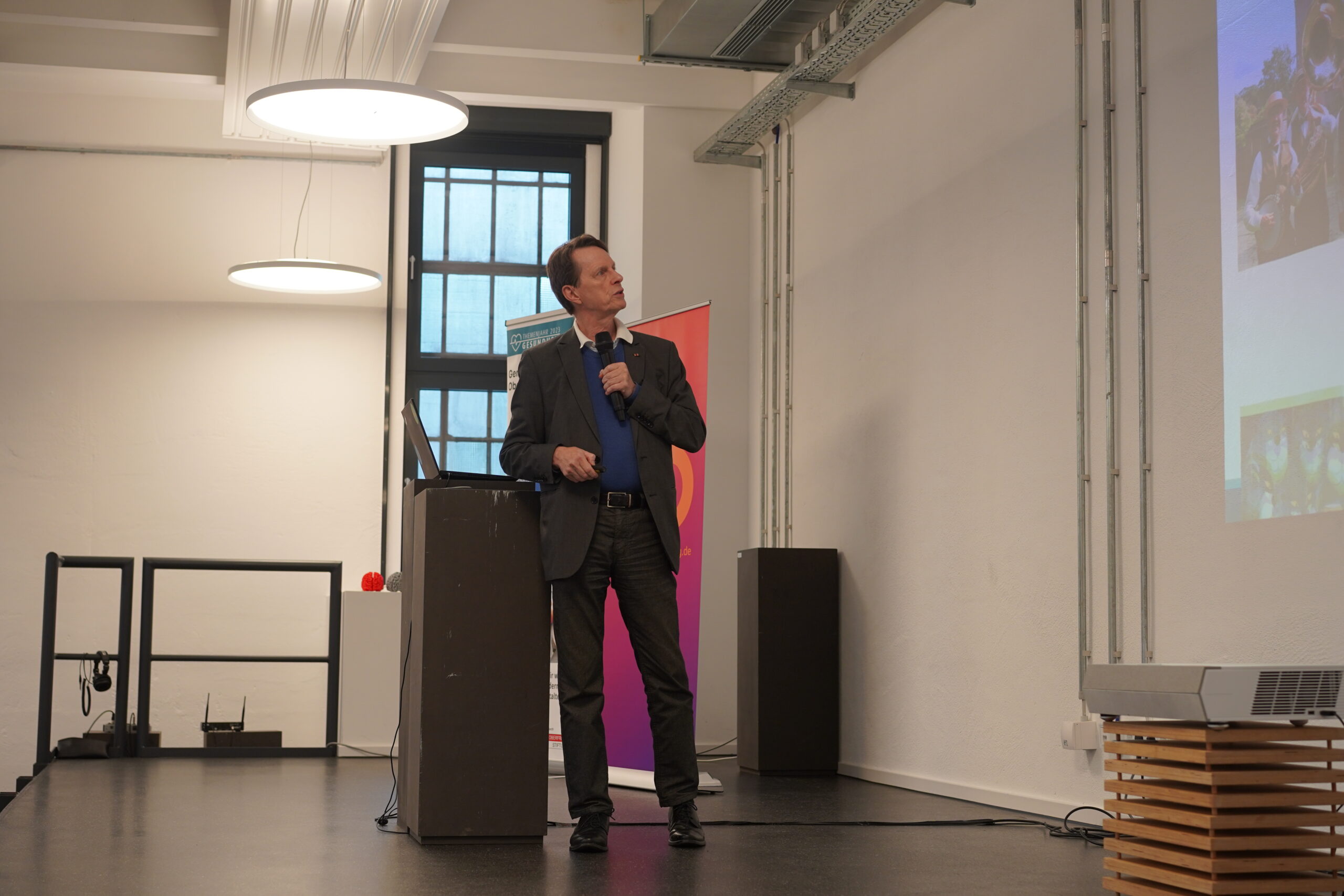 Eine Person im Anzug steht an einem Podium, spricht in ein Mikrofon und deutet auf eine Präsentationsfolie der Hochschule Coburg an einer Wand. Der Raum ist modern beleuchtet und verfügt über große Fenster.