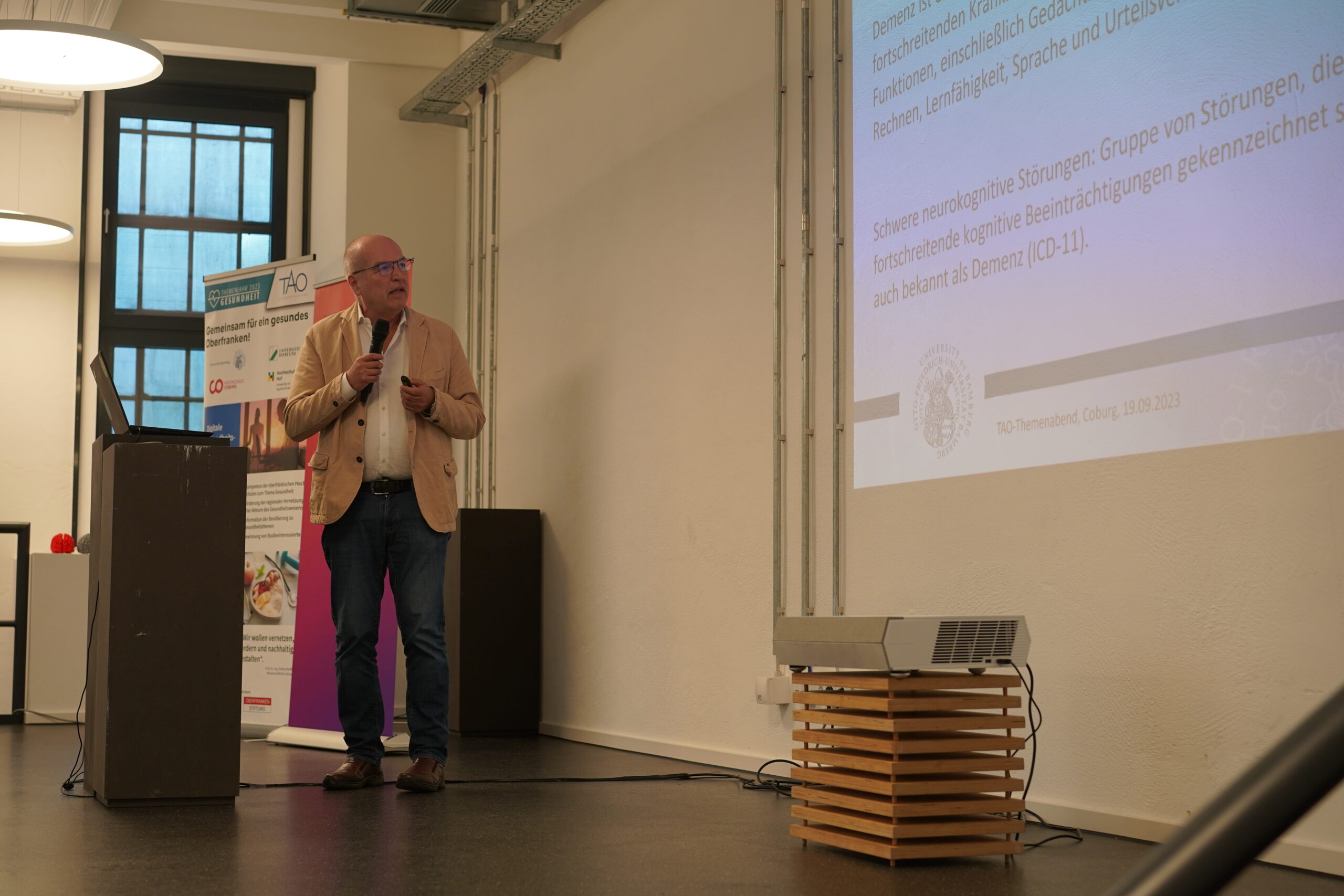 Ein Mann in einer beigen Jacke steht mit einem Mikrofon an einem Podium und präsentiert Folien in einem Raum mit einem Projektor an der Hochschule Coburg. Im Hintergrund sind ein Banner und ein Fenster zu sehen, die den akademischen Rahmen einrahmen.