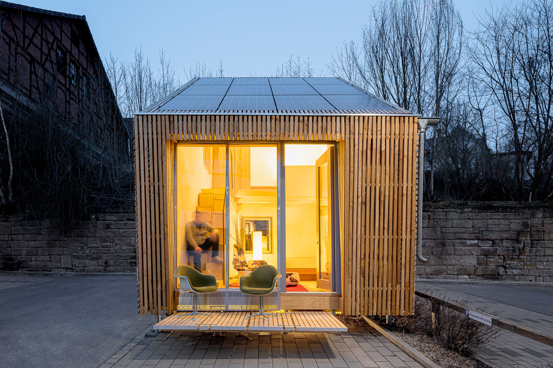 Ein kleines, modernes Holzhaus mit großen, von innen beleuchteten Fenstern, das ein warmes Interieur präsentiert, das an das Designethos der Hochschule Coburg erinnert. Vorne stehen zwei leere grüne Stühle auf der Veranda. Das Haus ist von kahlen Bäumen und Abendlicht umgeben.