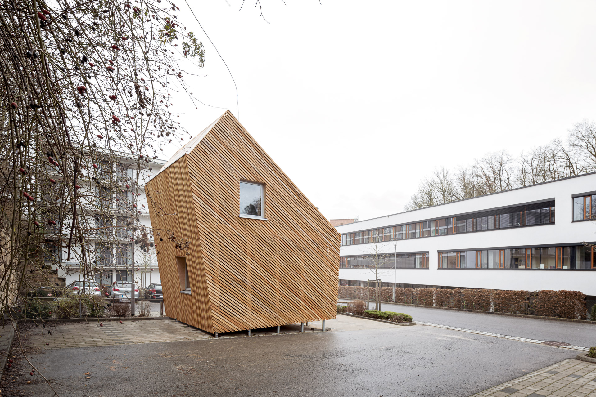 Ein kleines, einzigartig geformtes Holzgebäude mit schrägen Wänden steht in einer Straße in der Nähe der Hochschule Coburg. Es verfügt über ein großes Fenster und ist von modernen Gebäuden, Bäumen und einem wolkigen Himmel umgeben.