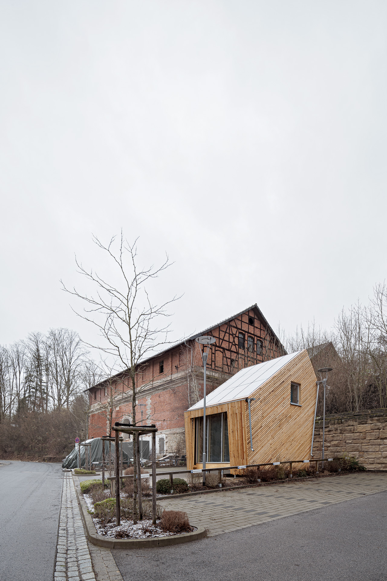 Ein modernes, kleines Holzhaus mit großen Fenstern steht am Straßenrand, neben einem älteren, rustikalen Backsteingebäude in der Nähe der Hochschule Coburg. Im Hintergrund sind kahle Bäume und ein bewölkter Himmel zu sehen, die eine kalte, winterliche Atmosphäre vermitteln.