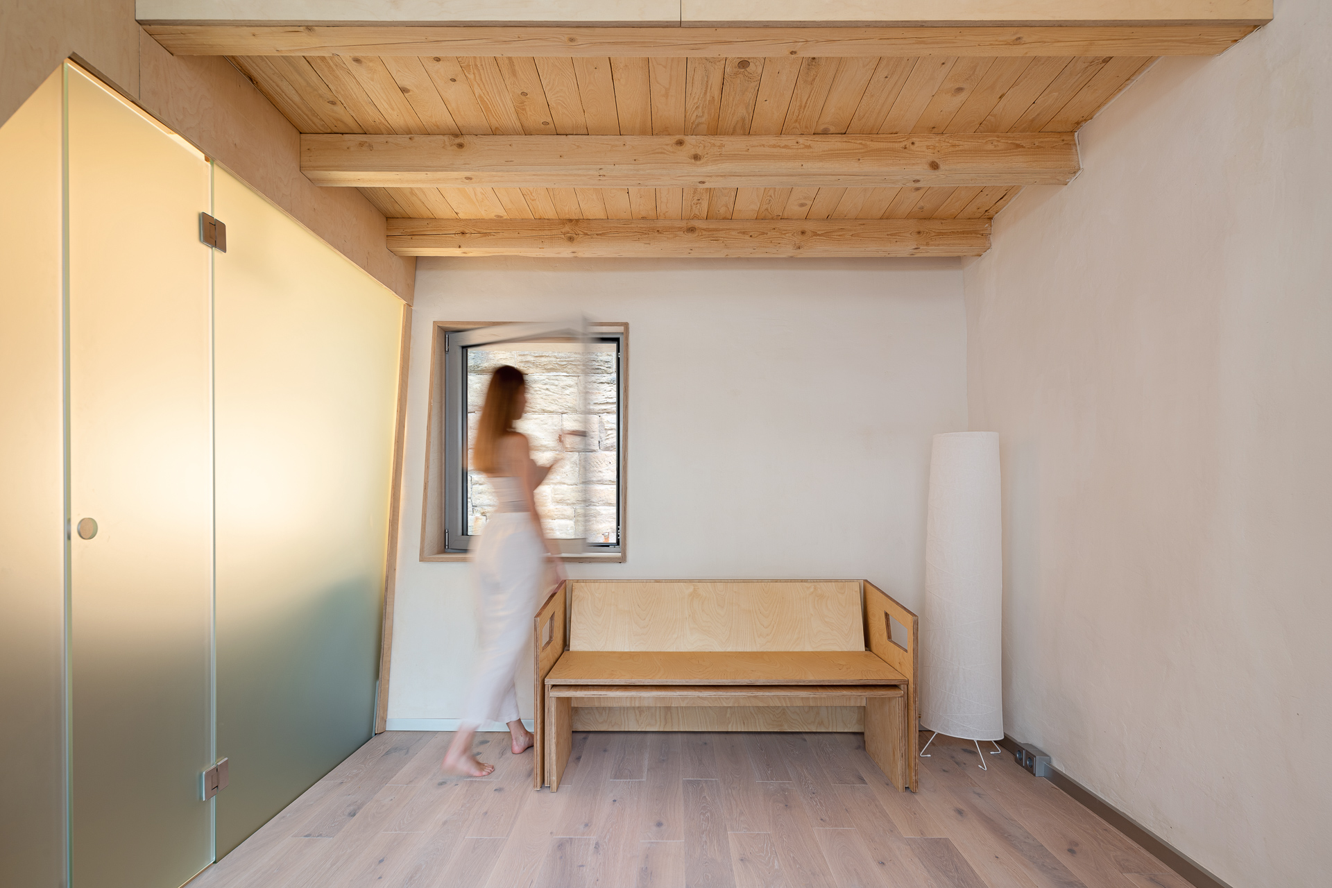 Eine verschwommene Frau steht neben einem Fenster in einem hellen, luftigen Raum der Hochschule Coburg mit Holzbalken an der Decke und einer Holzbank. Der Raum strahlt Bewegung aus und ist mit einer Milchglastür und einer großen, weißen zylindrischen Lampe ausgestattet.