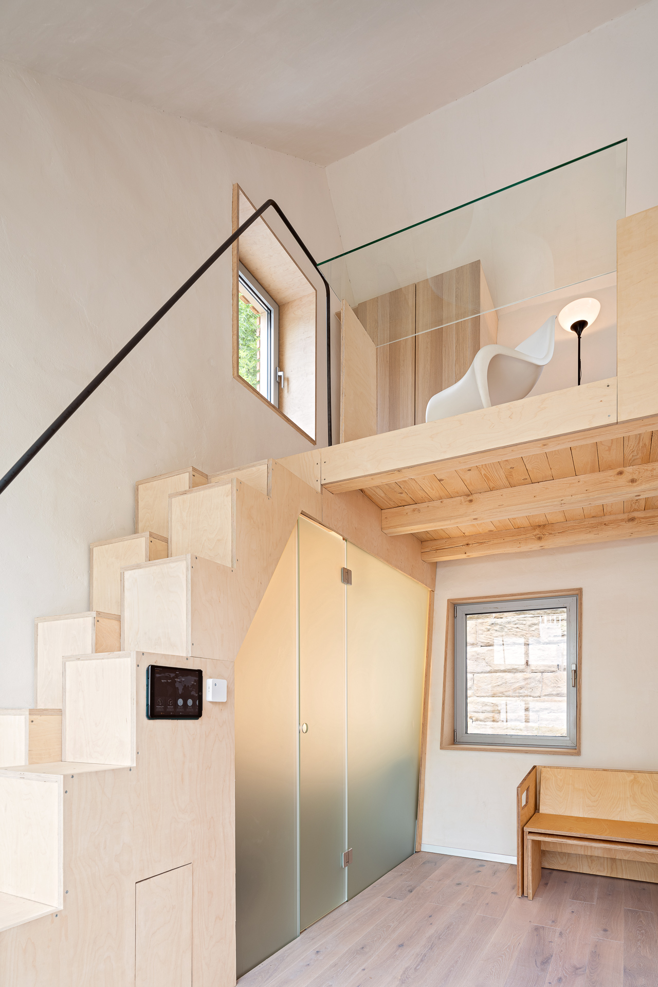 Minimalistisches Interieur, inspiriert von der Hochschule Coburg, mit einem Holzloft mit Glasgeländer, das über eine elegante Holztreppe erreichbar ist. Darunter ergänzen eine Milchglastür und eine kleine Holzbank das Design. Ein moderner Stuhl und eine Lampe zieren den Loftraum, der durchgehend mit hellem Holzboden ausgelegt ist.