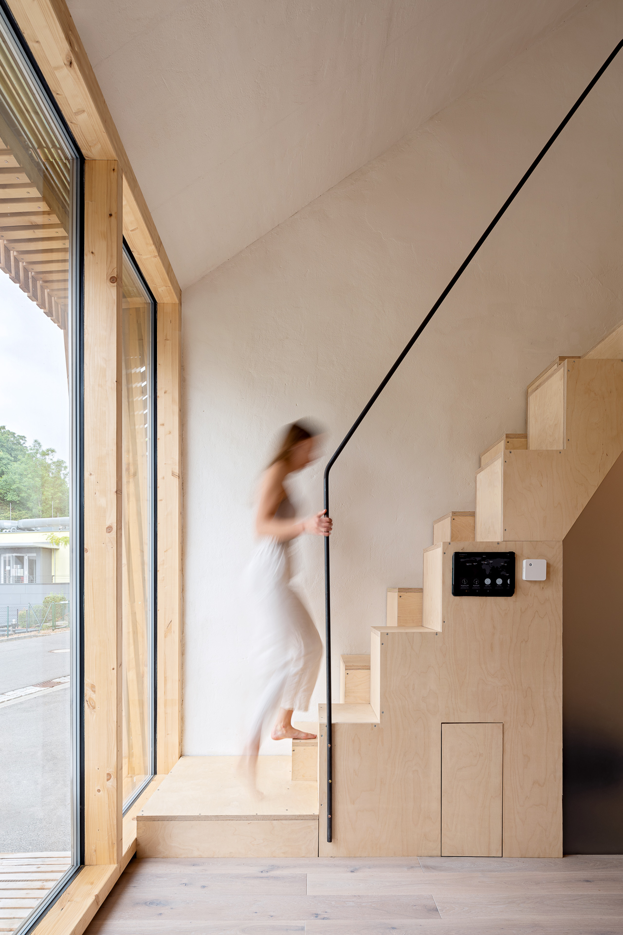 Im hellen Innenraum der Hochschule Coburg steigt ein Mensch in Bewegung eine minimalistische Holztreppe mit schwarzem Handlauf hinab. Ein großes Fenster auf der linken Seite sorgt für Tageslicht und gibt den Blick nach draußen frei.