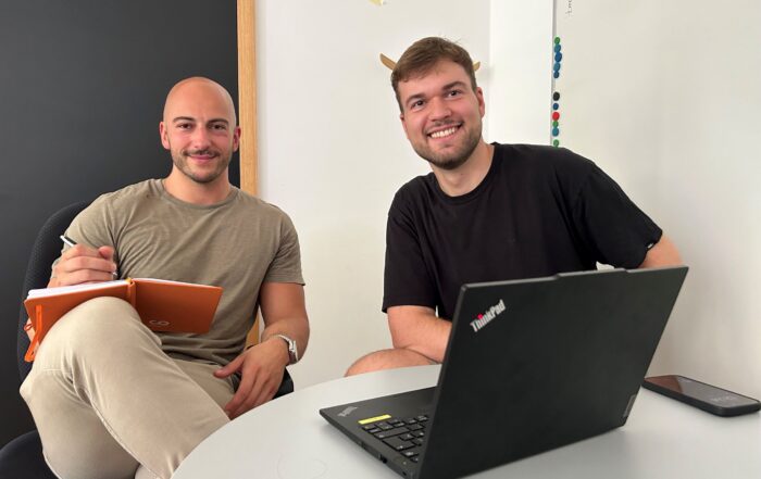 Two people sitting at a table, one holding an orange notebook and pen, the other using a laptop. They're smiling at the camera. The background features a whiteboard with some writing and a black door, hinting at their collaborative project for Hochschule Coburg.