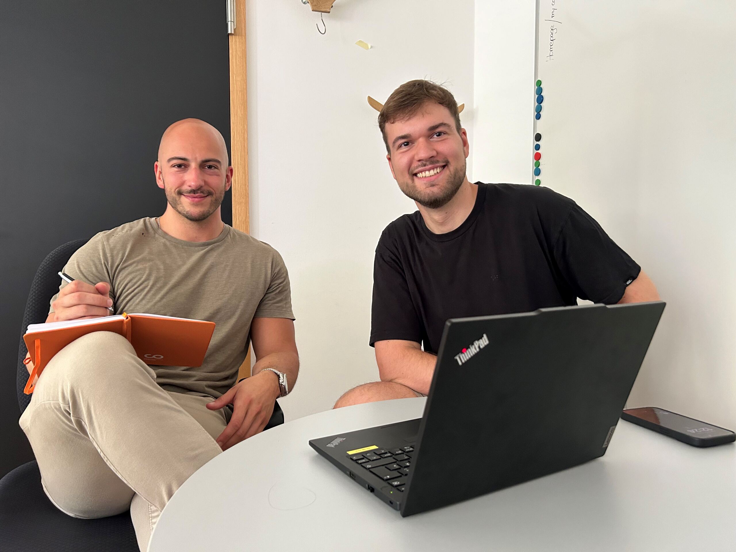 Zwei Männer sitzen an einem runden Tisch in einem Besprechungsraum der Hochschule Coburg. Einer hält ein orangefarbenes Notizbuch und einen Stift in der Hand, während der andere einen Laptop benutzt. Beide lächeln und sind entspannt, ihr Telefon liegt auf dem Tisch. Ein Whiteboard und eine dunkle Wand dienen als Hintergrund für ihre gemeinsame Sitzung.