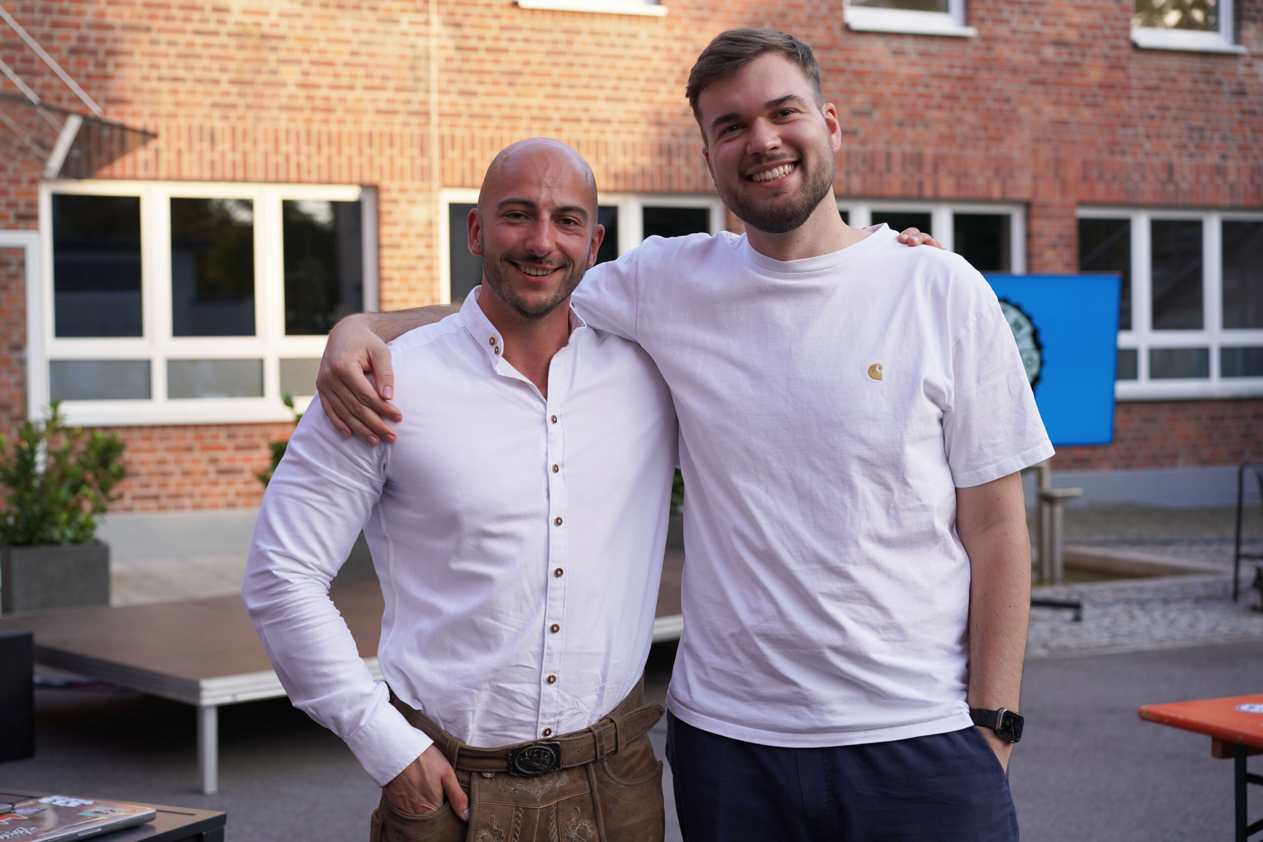 Two men stand close together outside, smiling at the camera. The man on the left wears a white button-up shirt and brown pants, while the one on the right is in a white T-shirt and dark pants. They're in front of a brick building with windows, possibly part of Hochschule Coburg.