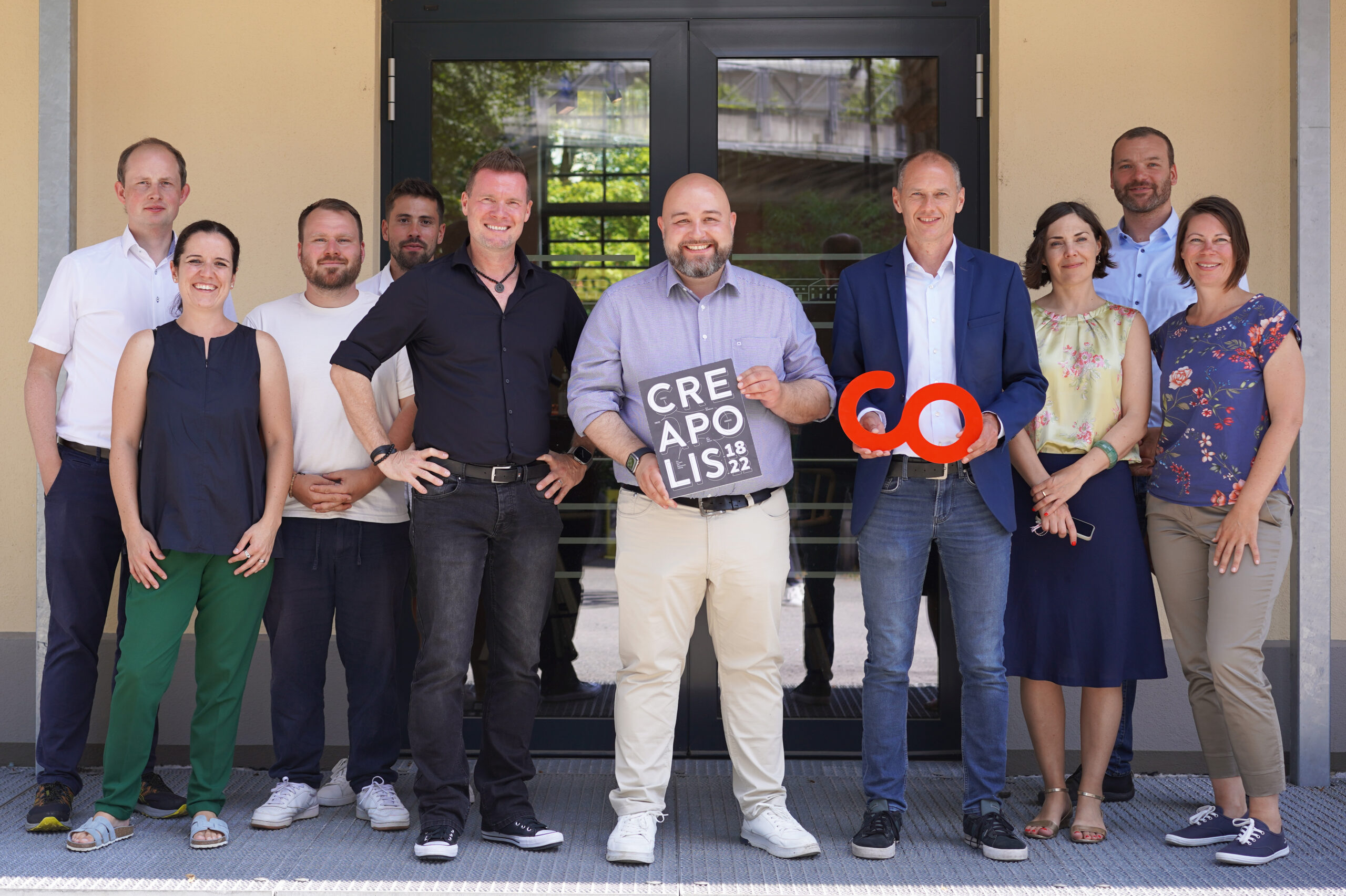 A group of ten people, standing and smiling in front of a building's entrance at Hochschule Coburg. One person holds a small sign with "CREAPOLIS 18/12," while another holds a red "60" sculpture. They are dressed in a mix of casual and business attire.