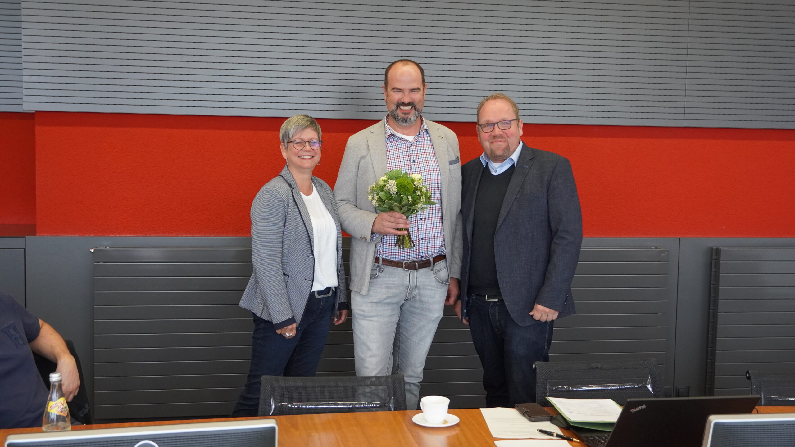 Drei Menschen stehen in einem Besprechungsraum der Hochschule Coburg. Die Person in der Mitte hält einen Blumenstrauß in der Hand. Alle lächeln, vor ihnen auf dem Tisch stehen Laptops und eine Kaffeetasse.