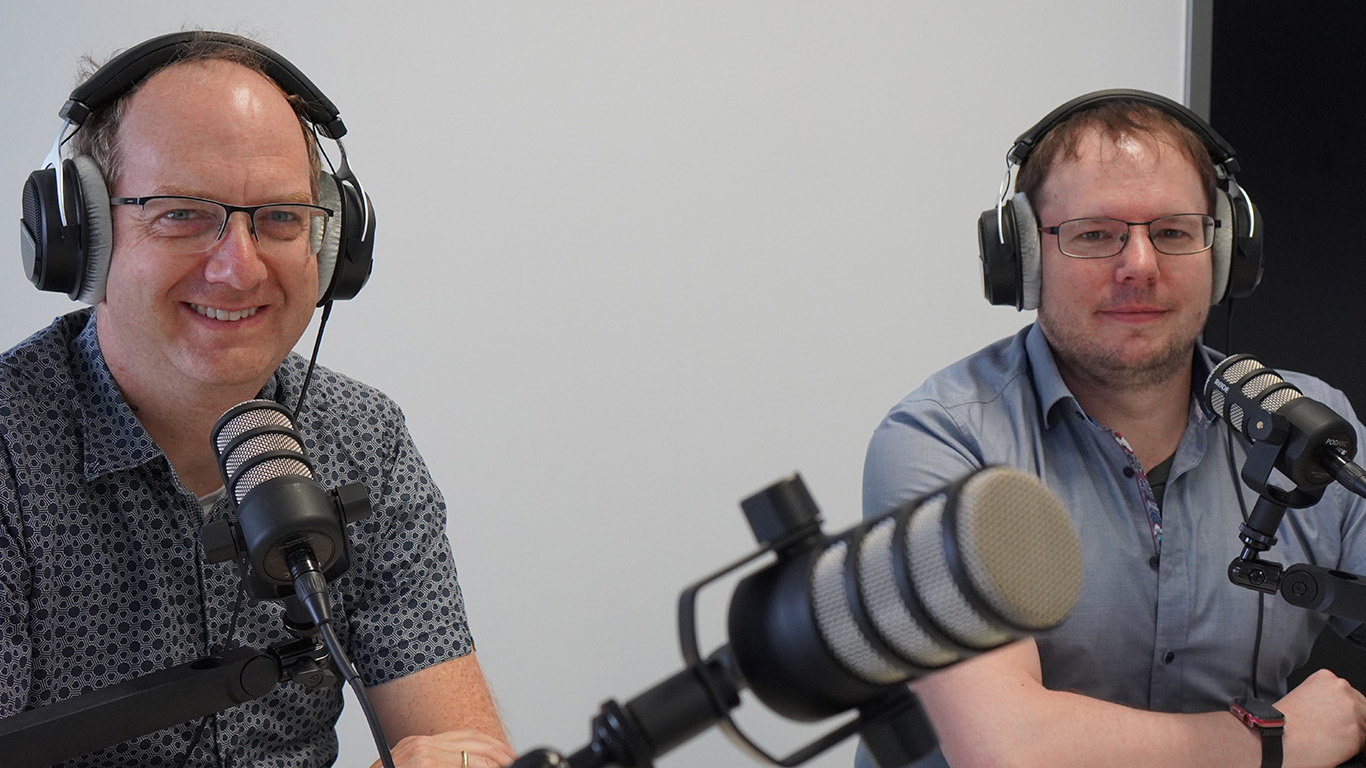 Two people sit at a table in front of microphones, wearing headphones and smiling as they likely record a podcast or radio show. The plain white wall offers a neutral backdrop, perhaps suggesting they're broadcasting from Hochschule Coburg's dedicated media space.