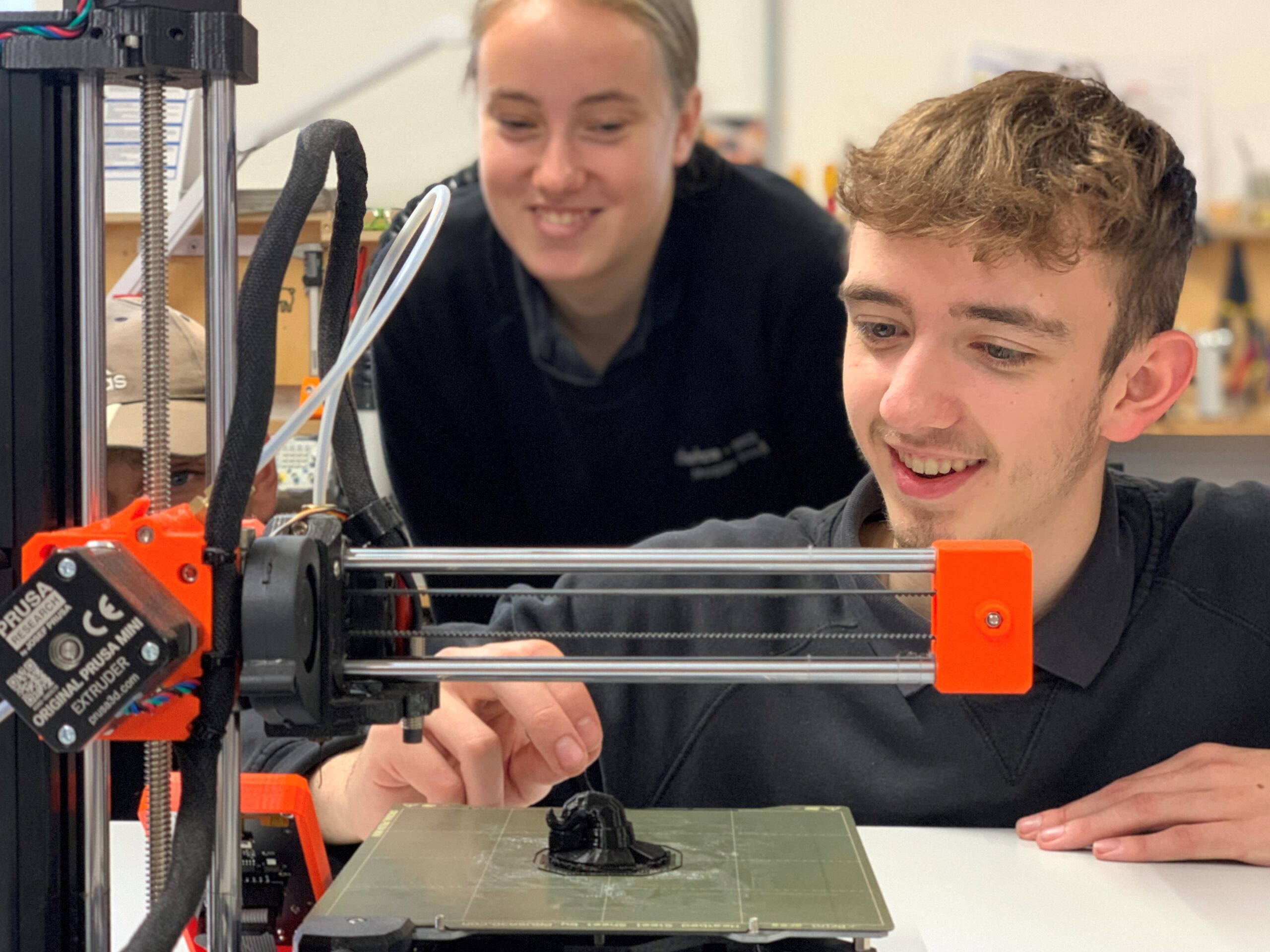 Zwei Personen konzentrieren sich auf einen 3D-Drucker, der an der Hochschule Coburg in Betrieb ist. Die Person im Vordergrund justiert ihn, während die Person im Hintergrund zusieht. Der teilweise gebaute Drucker druckt aktiv ein kleines schwarzes Objekt auf der Plattform.