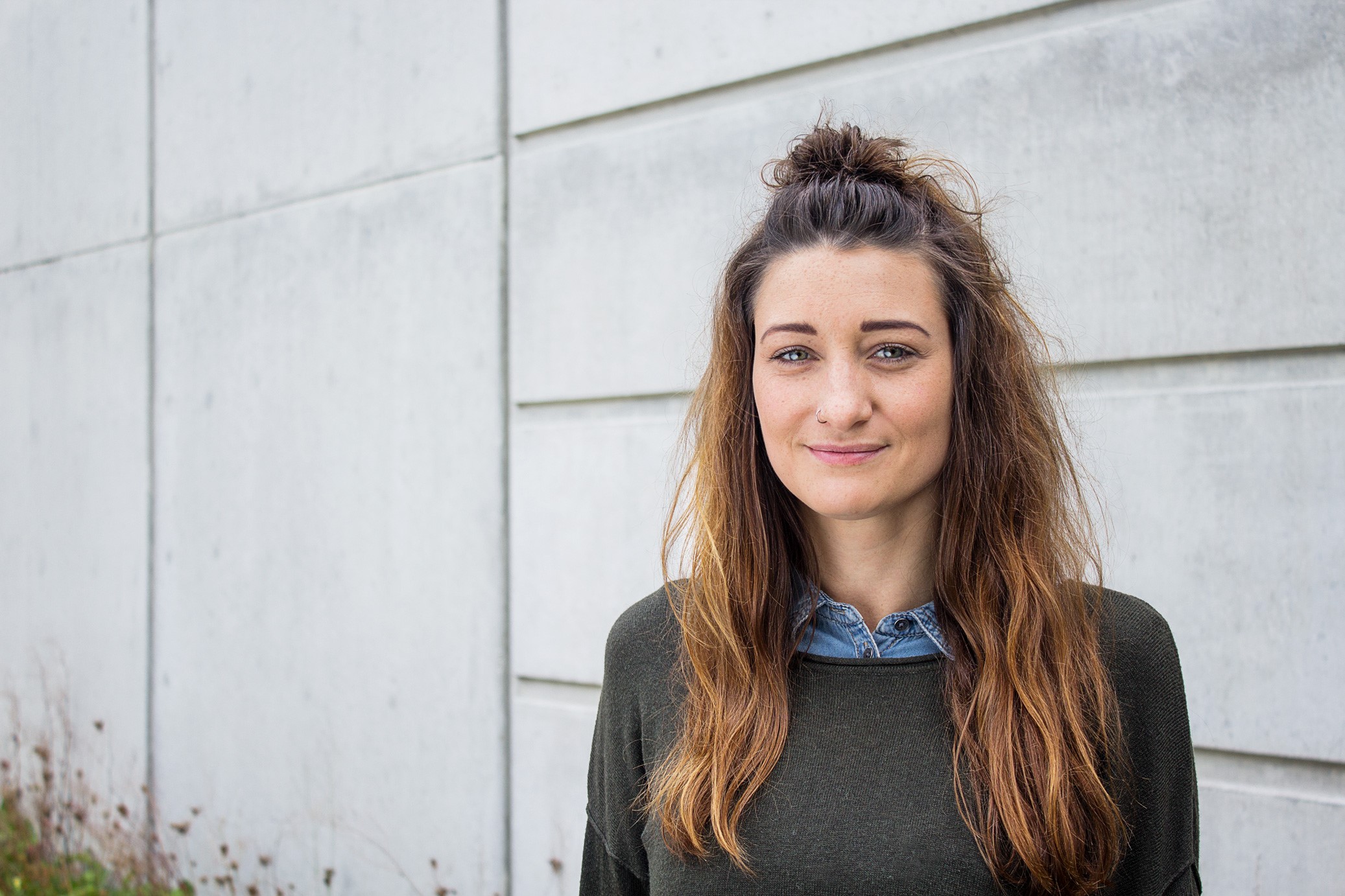 Eine Frau mit langen braunen Haaren, die halb hochgesteckt sind, steht an einer Betonwand der Hochschule Coburg. Sie trägt einen grünen Pullover über einem Jeanshemd und lächelt in die Kamera. Am unteren Ende der Wand ist Gras zu sehen.