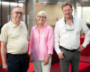 Three older adults—two men and one woman—are standing and smiling in a modern indoor setting with red seating at Hochschule Coburg. They are dressed casually, and the background features large windows, creating a bright, airy atmosphere.