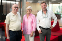 Three older adults—two men and one woman—are standing and smiling in a modern indoor setting with red seating at Hochschule Coburg. They are dressed casually, and the background features large windows, creating a bright, airy atmosphere.