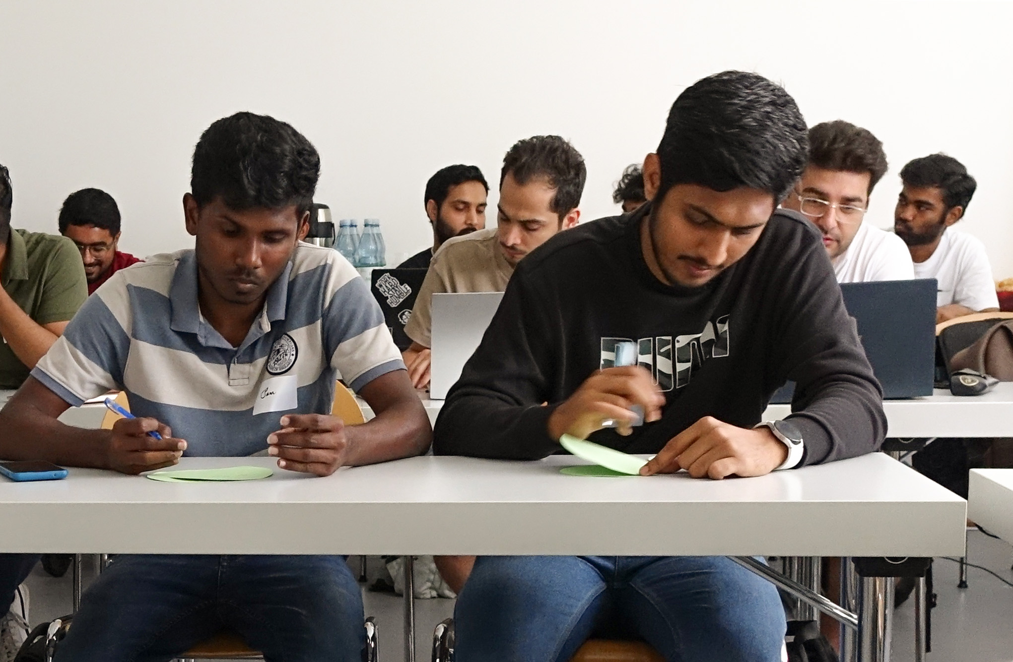 A group of people seated at desks, immersed in writing on paper, appears to be in a classroom setting at Hochschule Coburg. They focus intently on their tasks, with some having laptops open in front of them.