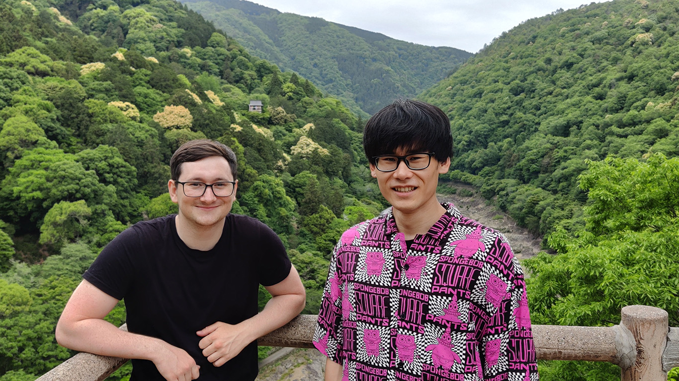 Two people stand on a wooden railing with lush green mountains in the background, capturing the essence of Hochschule Coburg's natural appeal. The person on the left wears a black shirt, and the one on the right sports a pink patterned shirt. The scene is bright and serene.