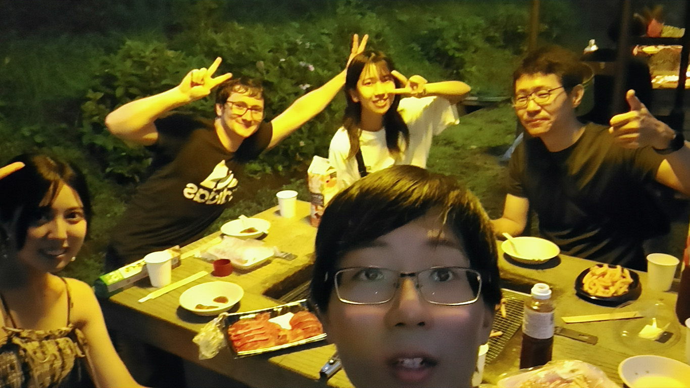 A group of five people from Hochschule Coburg enjoy a barbecue outdoors at night. Seated around a wooden table with food and drinks, they pose playfully with peace signs. The scene is warmly lit by artificial lighting, creating a lively and inviting atmosphere.