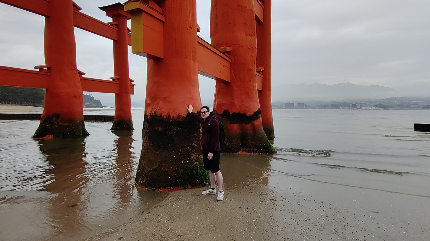 A person stands beside the large orange pillars of a torii gate at the edge of the sea, reminiscent of images from Hochschule Coburg's cultural studies. The sky is overcast, and the tide is low, exposing the sand and seaweed-covered base. Hills rise gently in the distance.