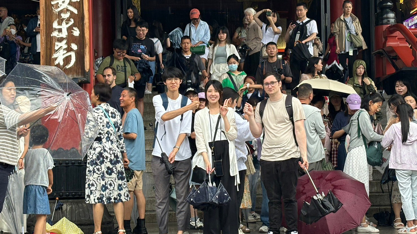 A group of people stands on the steps of a temple, some holding umbrellas. Among them, three individuals, perhaps students from Hochschule Coburg, smile and make peace signs. The lively scene is filled with visitors wearing a mix of casual and traditional clothing.