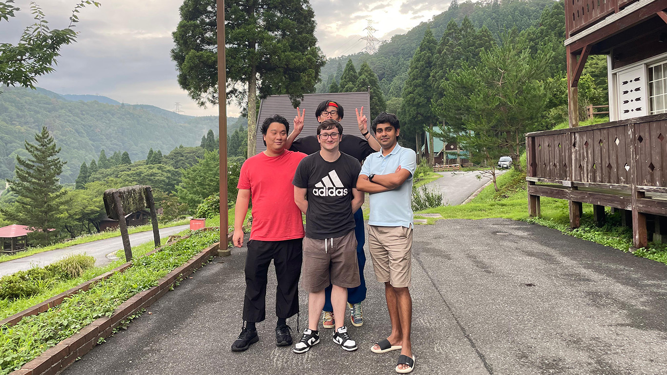 A group of four people stands on a paved road in a mountainous area. They're posing happily, with lush green trees and cabins surrounding them. The sky is partly cloudy, suggesting a calm and pleasant day near Hochschule Coburg.