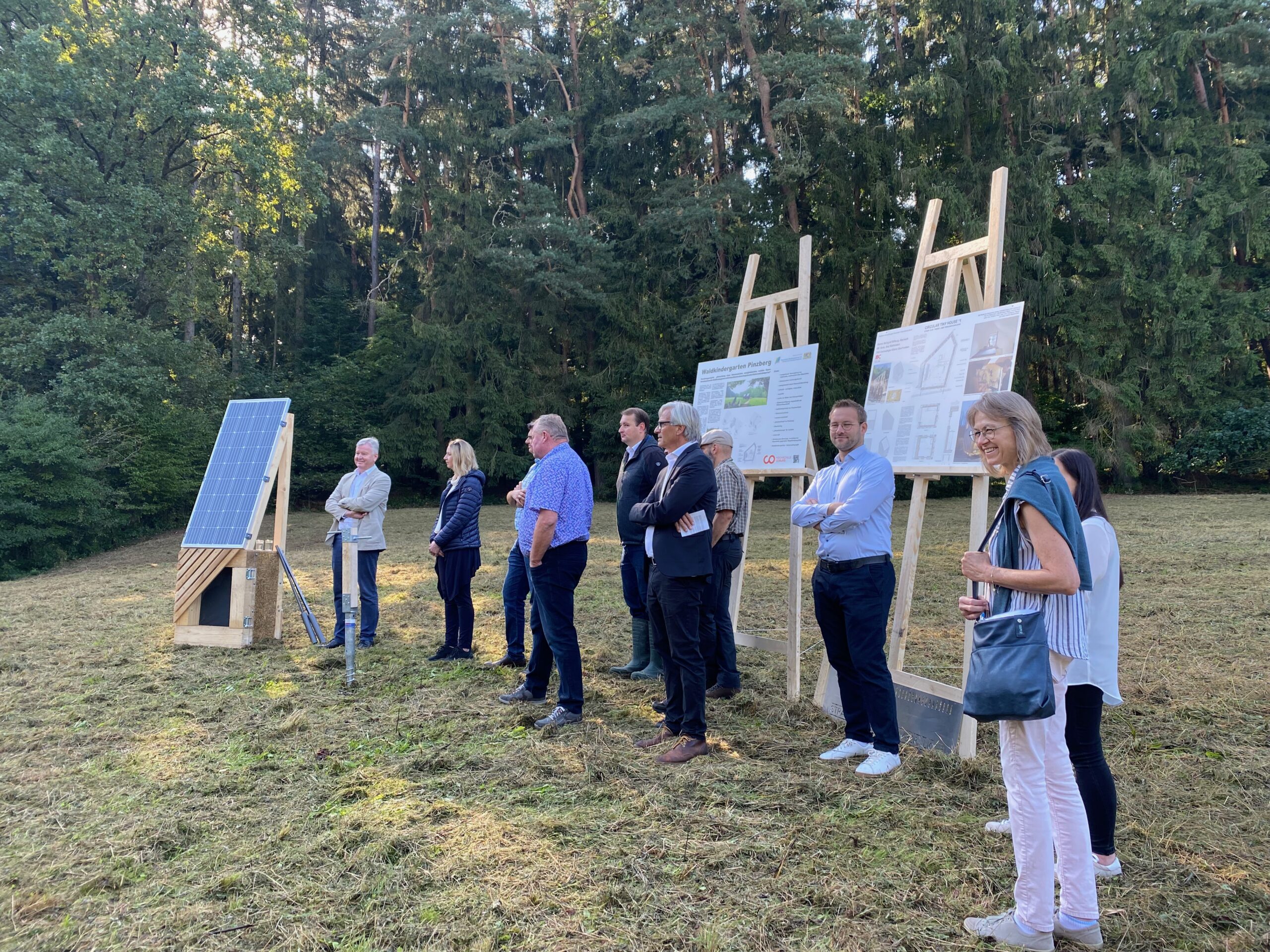 Eine Gruppe von Mitarbeitern der Hochschule Coburg steht im Freien auf einer Wiese neben einer Solarpanel-Ausstellung und großen Informationsplakaten auf Staffeleien. Im Hintergrund sind hohe Bäume zu sehen. Einige beobachten die Präsentation, während andere Mitarbeiter der Hochschule Coburg an der Diskussion teilnehmen.