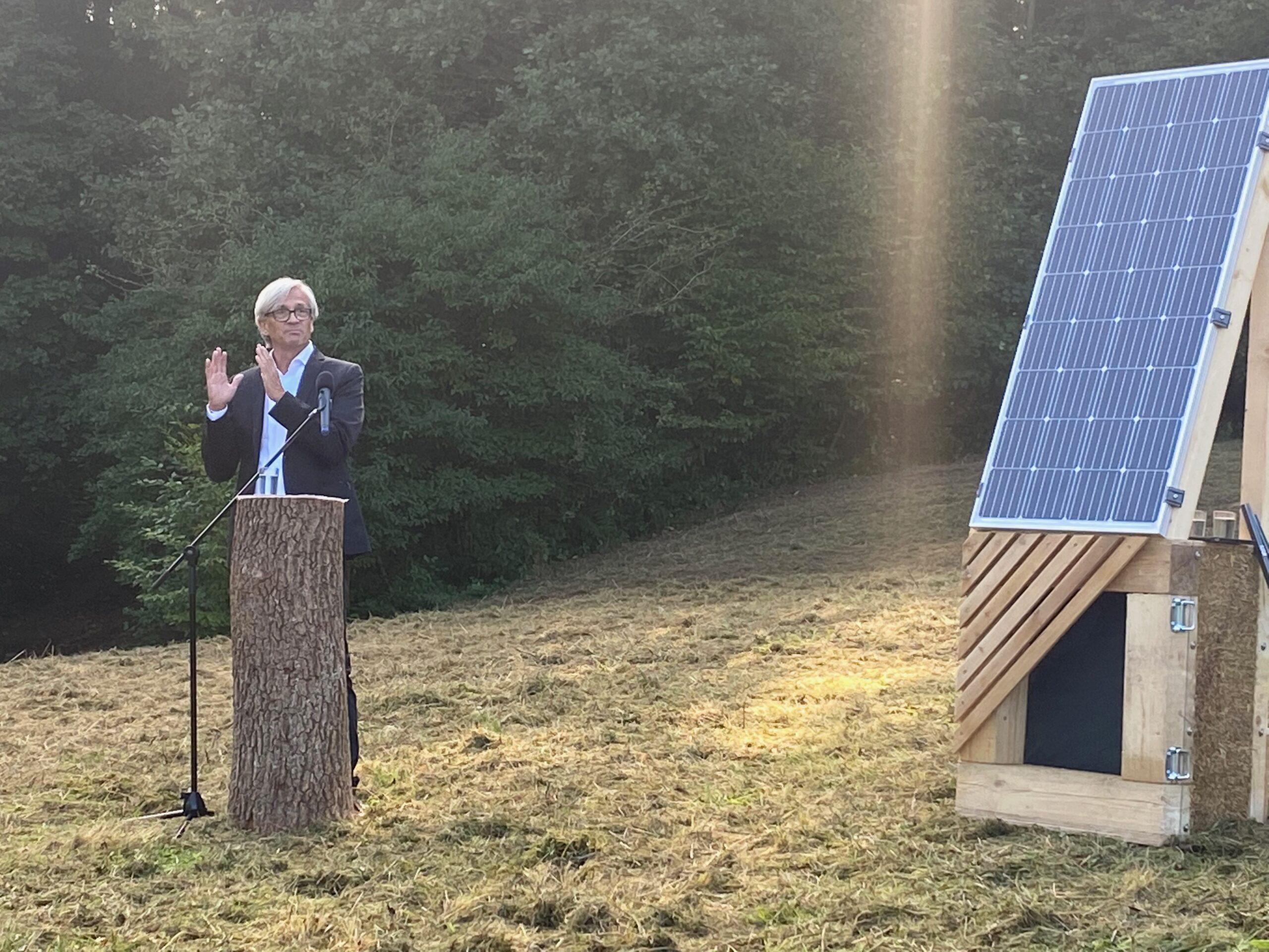 Eine Person der Hochschule Coburg spricht an einem Außenpodium aus einem Baumstumpf, rechts davon steht ein Solarpanel auf einem Holzständer. Sie stehen auf einer von Bäumen umgebenen Rasenfläche, während Sonnenlicht durch die Szene strömt.