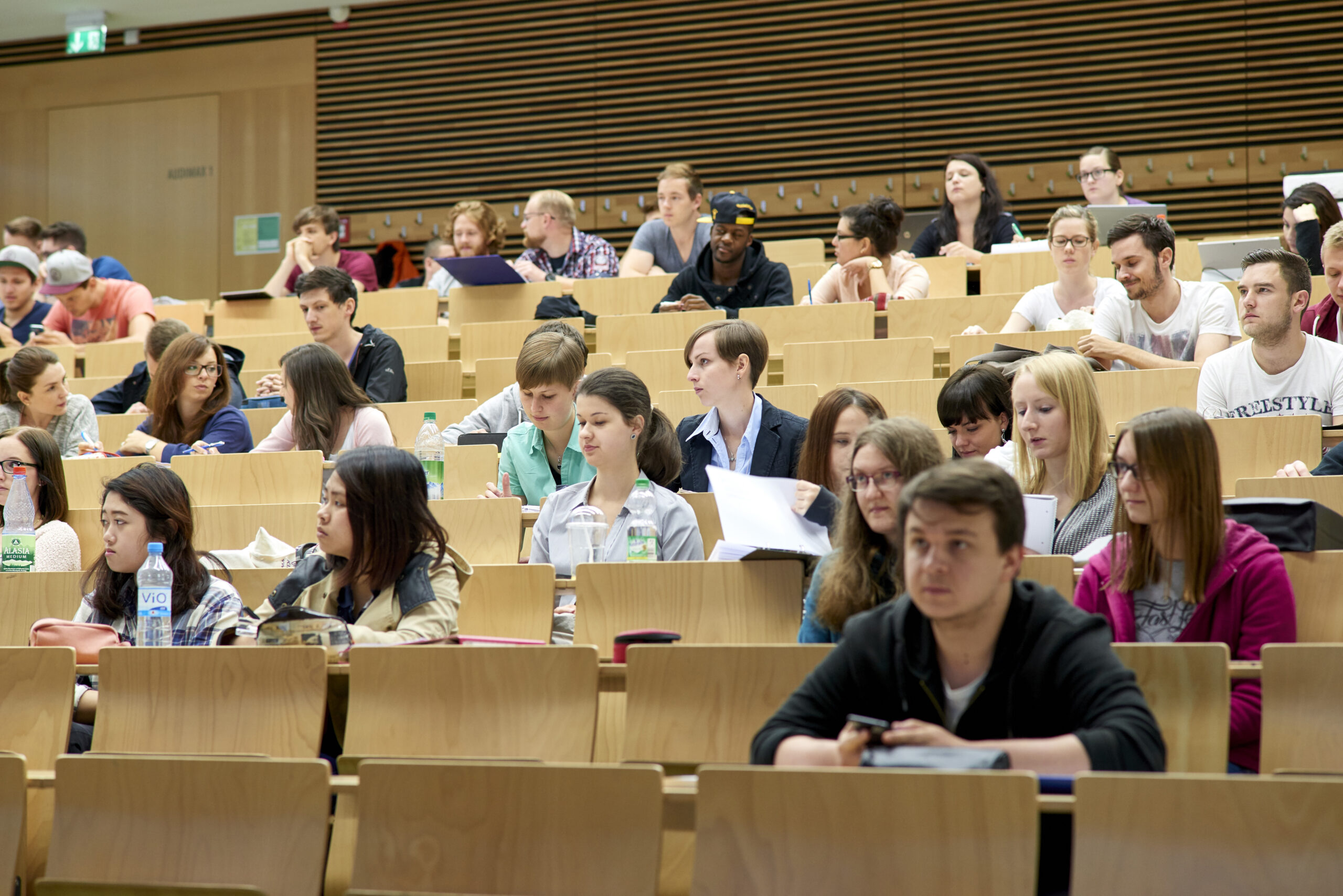 Im Hörsaal der Hochschule Coburg hört eine bunt gemischte Gruppe von Studierenden aufmerksam zu und macht sich Notizen. Der Raum ist mit Holzbänken und neutralen Farben gestaltet. Manche Studierende haben Laptops, andere schreiben in Notizbüchern.