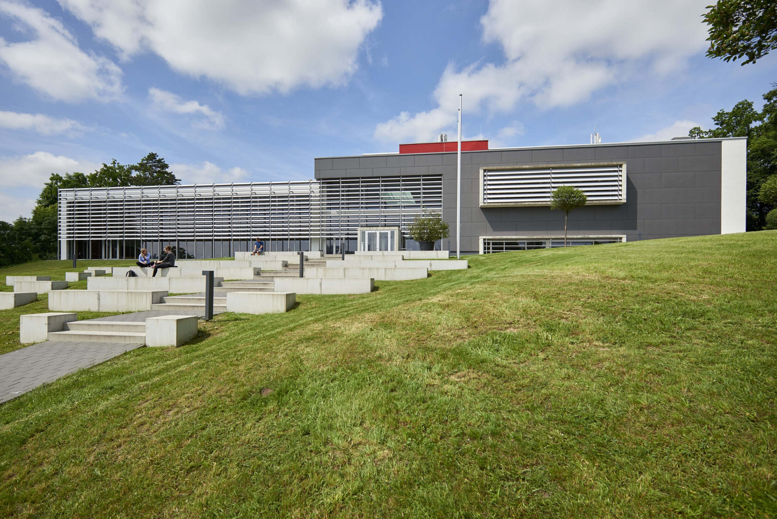 Ein modernes Gebäude, das möglicherweise an die Architektur der Hochschule Coburg erinnert, verfügt über eine graue Fassade mit weißen Lamellenfenstern auf einem Grashang. Betonstufen im Vordergrund bieten Sitzgelegenheiten. Der Himmel ist größtenteils klar mit ein paar Wolken, und ganz rechts sind Bäume zu sehen.