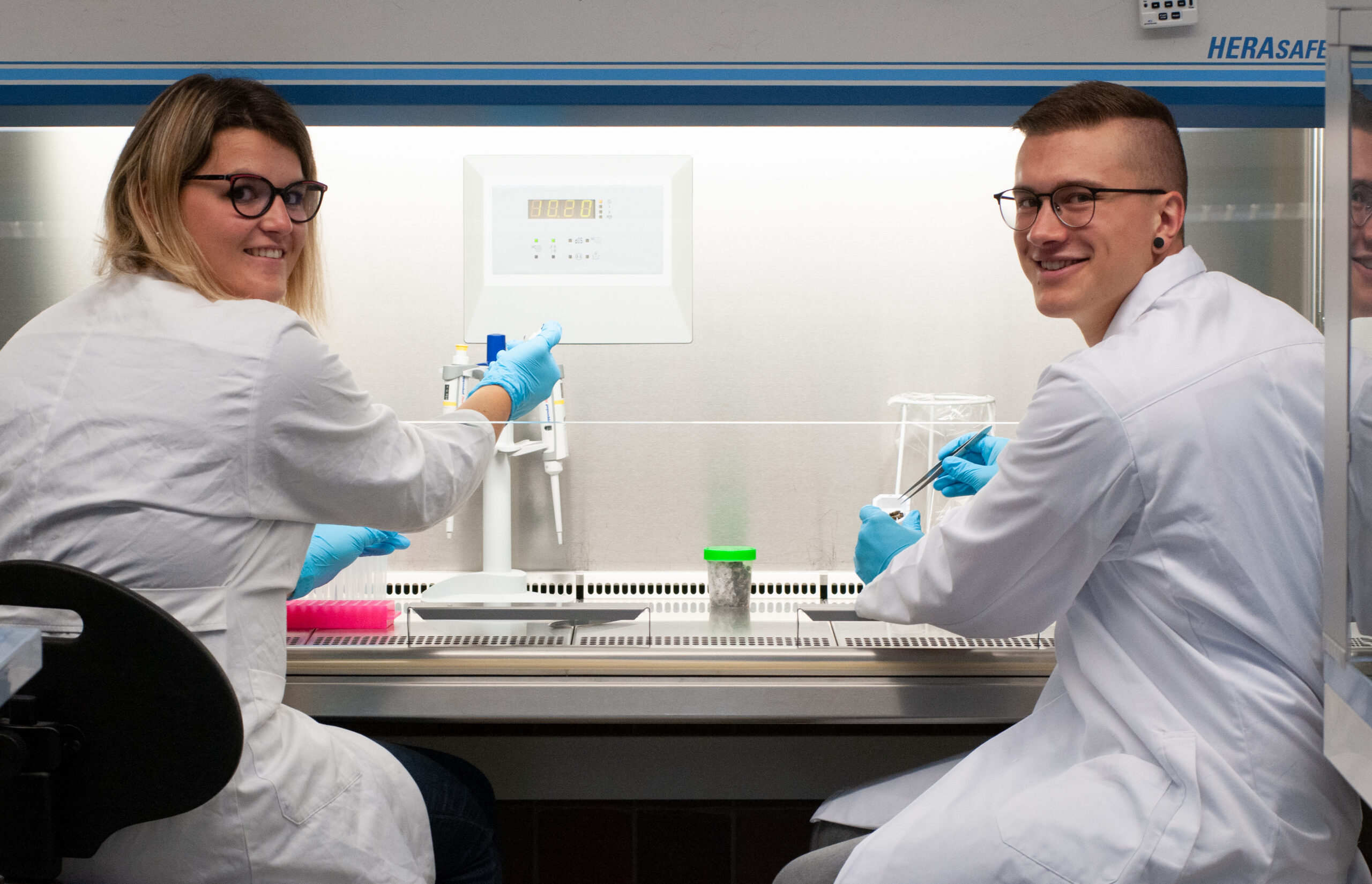 Zwei Wissenschaftler der Hochschule Coburg arbeiten in weißen Kitteln und blauen Handschuhen in einem Labor. Sie sitzen an einer Sicherheitswerkbank und lächeln inmitten verschiedener Laborgeräte in die Kamera.