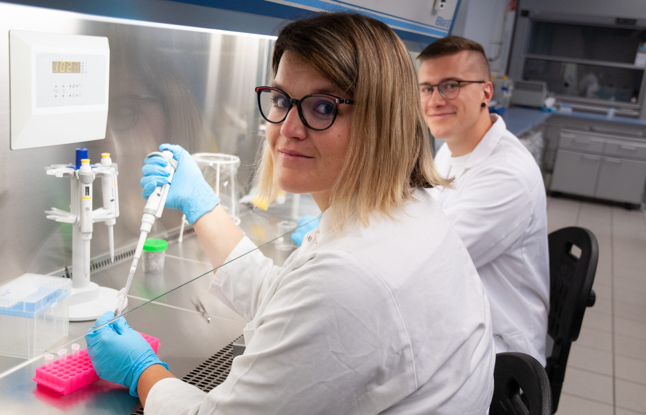 In einem Labor der Hochschule Coburg sind zwei Wissenschaftler in Laborkitteln fleißig bei der Arbeit. Die Frau vorn sitzt mit blauen Handschuhen und Brille an ihrem Arbeitsplatz und benutzt eine Pipette. Hinter ihr sitzt der Mann, ebenfalls mit Brille, und lächelt, umgeben von einer Reihe von Laborgeräten.