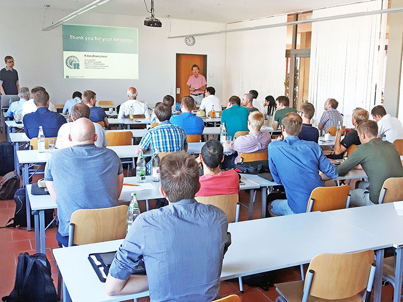 Der Hörsaal der Hochschule Coburg ist voller Menschen, die an Tischen sitzen und aufmerksam auf einen Bildschirm blicken, auf dem eine Präsentation läuft. Der Vortragende steht vorne, und auf den Tischen sind verschiedene Flaschen und Papiere zu sehen.