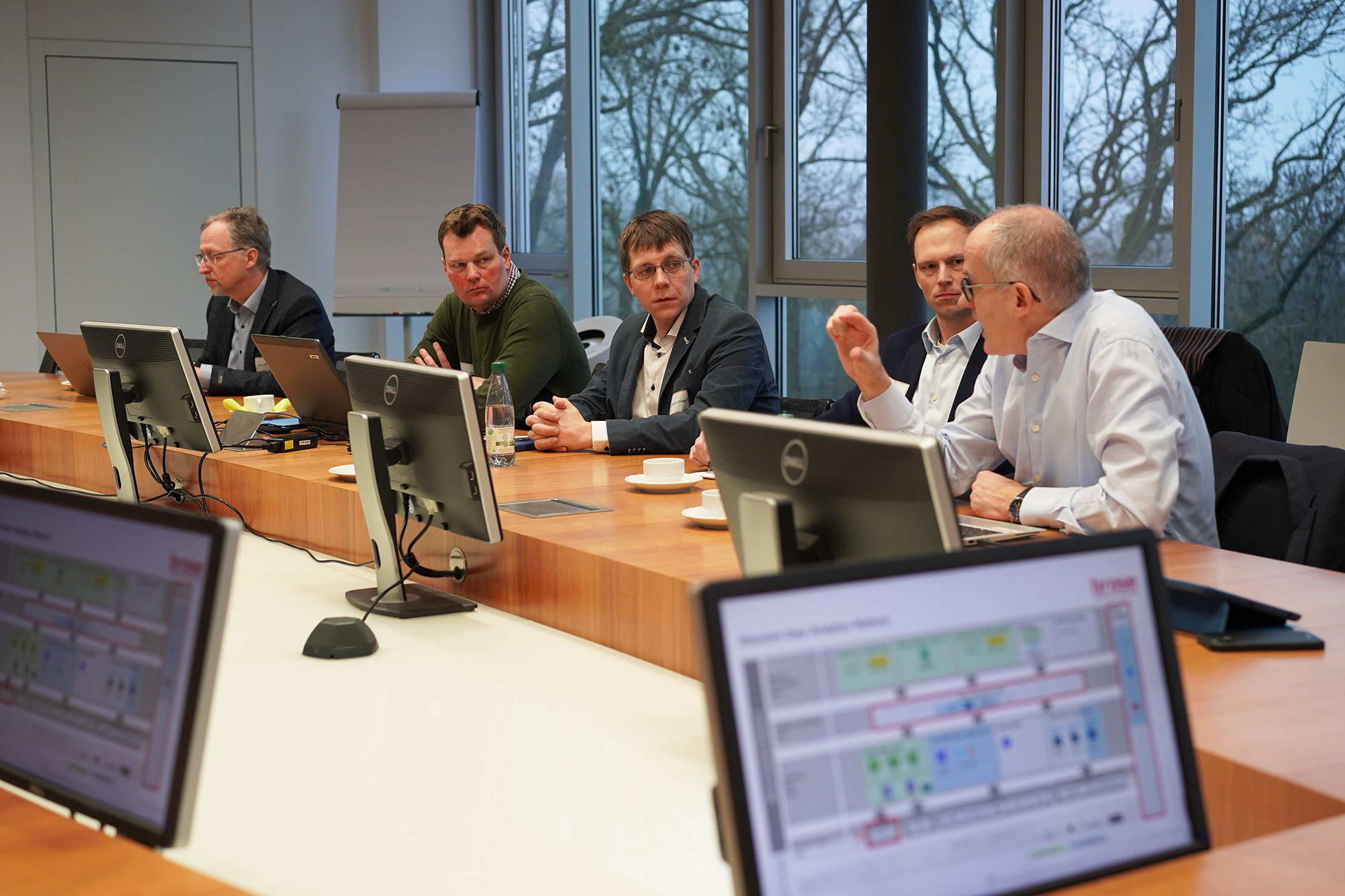 A group of five men are seated around a conference table with computers in front of them at Hochschule Coburg. One man gestures as they discuss something. Large windows in the background reveal trees outside. The atmosphere appears professional and focused.