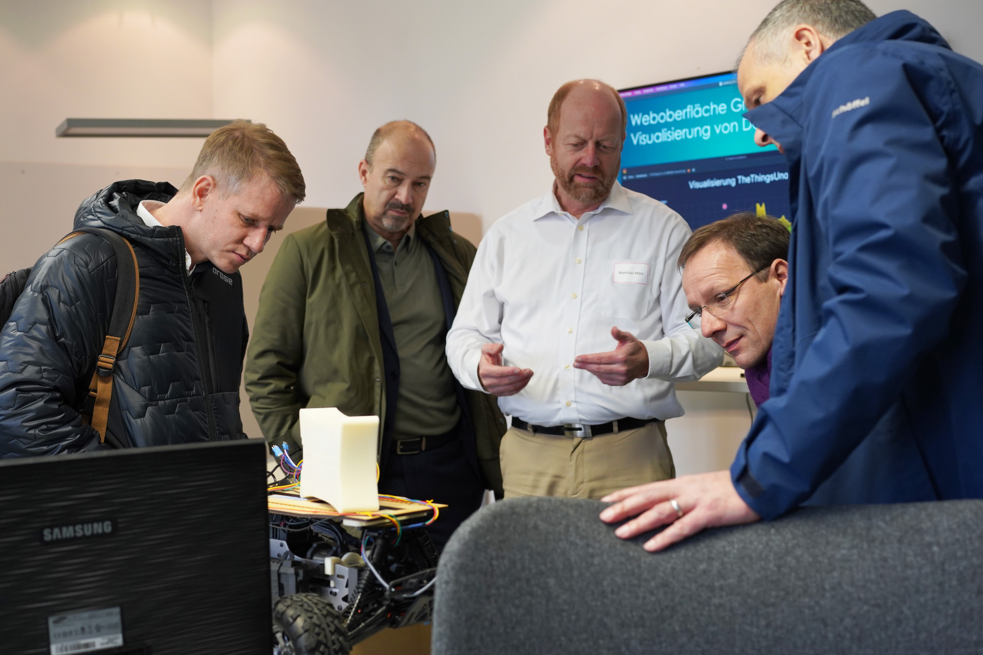 Four men gather around a table, attentively examining a piece of technology. One speaks while others listen closely. A Samsung monitor and technical equipment are visible. A sign with German text on the wall hints at their location in Hochschule Coburg, fostering innovation and collaboration.