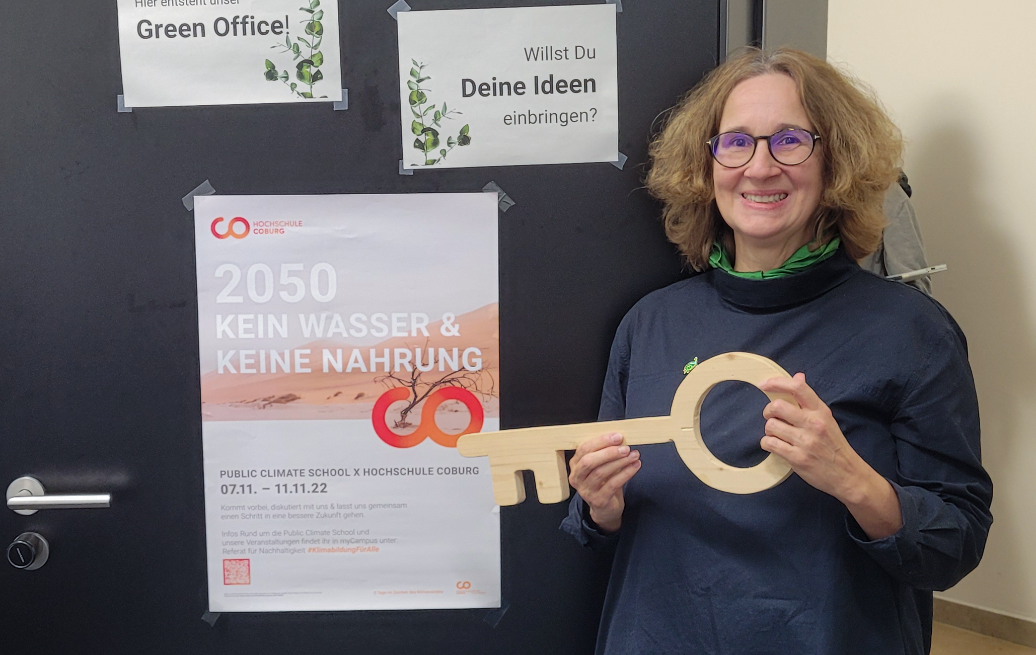 Eine Person steht lächelnd neben einer Tür und hält einen großen Holzschlüssel in der Hand. An der Tür hängt ein Plakat über eine öffentliche Klimaschulveranstaltung an der Hochschule Coburg und ein Schild, auf dem um Ideen gebeten wird. Die Person trägt eine Brille und ein dunkles Outfit.