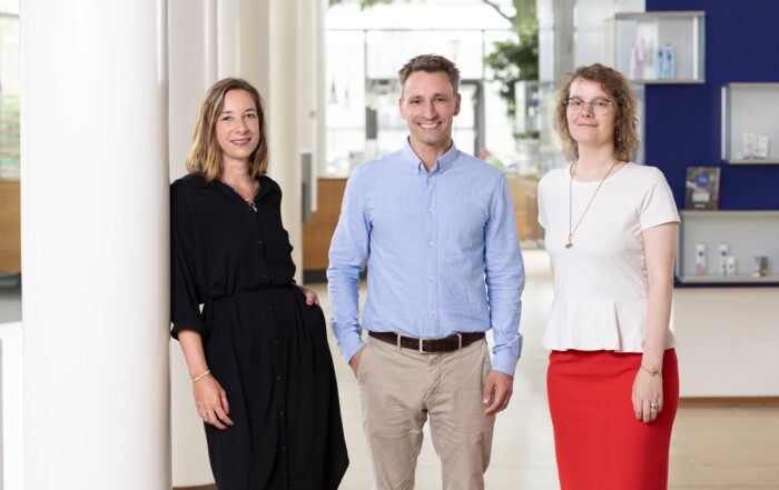 Three people stand indoors in a well-lit, modern space at Hochschule Coburg. A woman in a black dress leans against a white column, while a man in a blue shirt and beige pants stands in the center. Another woman, wearing a white top and red skirt, is positioned on the right.